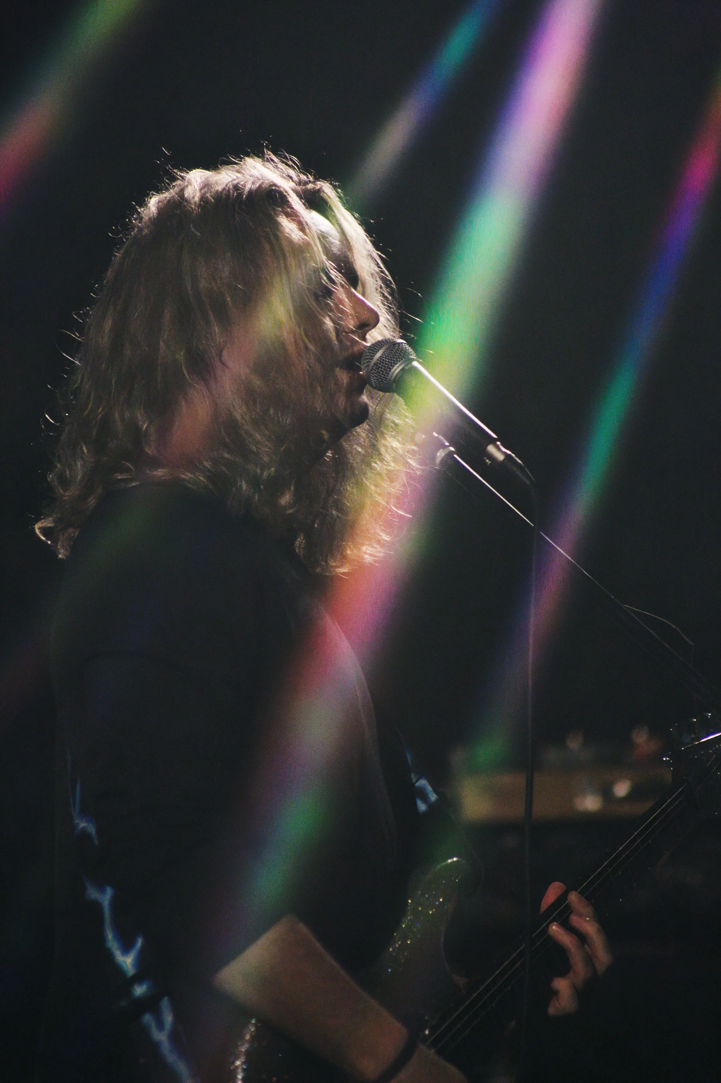 A musician with shoulder-length curly hair singing into a microphone while playing an electric guitar, with rainbow-colored light streaks across a dark background.