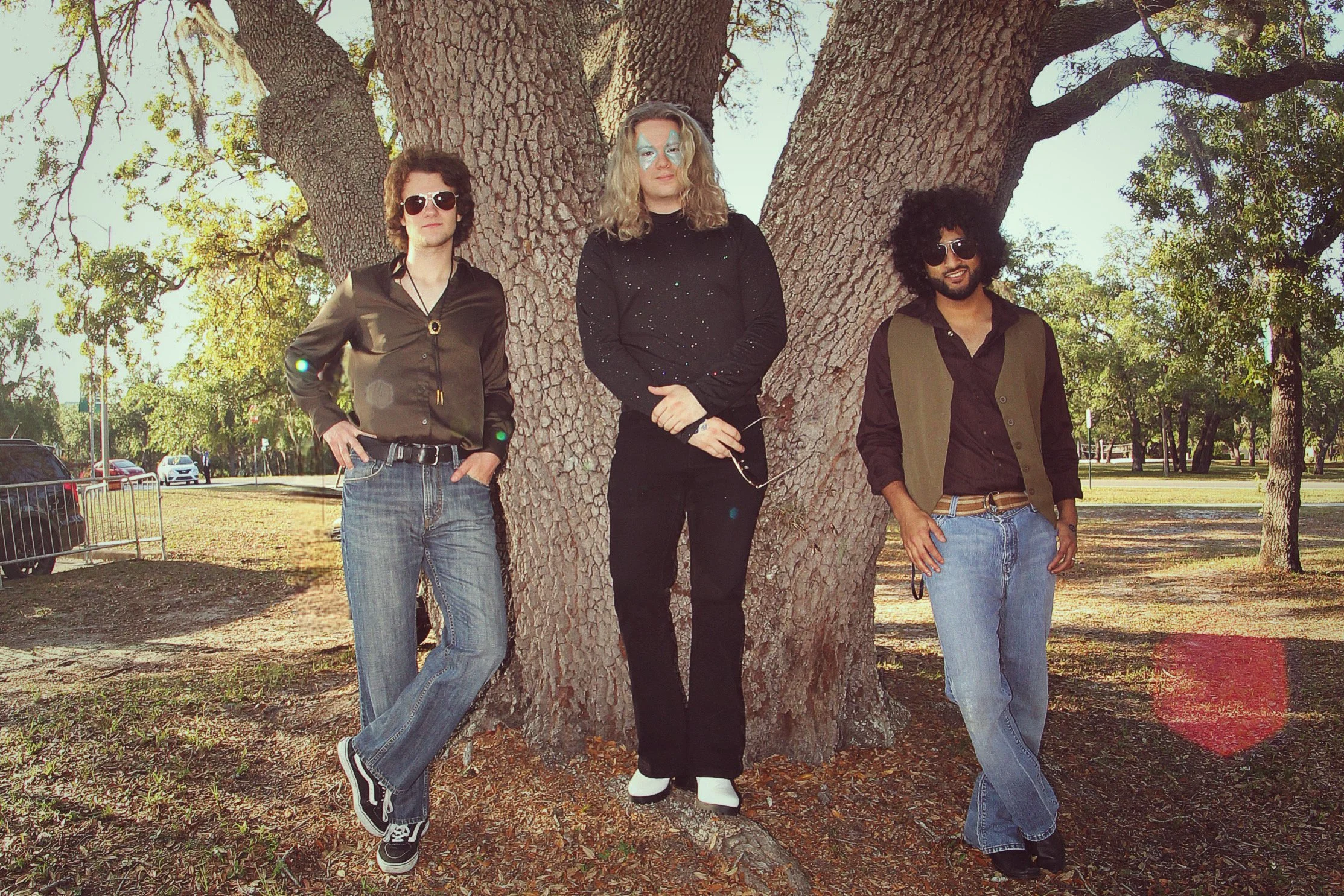 Three people standing outdoors in front of a large tree on a sunny day. The person on the left is wearing sunglasses, a black satin shirt, jeans, and sneakers. The person in the middle is wearing makeup, a black sweater with sparkles, black pants, an