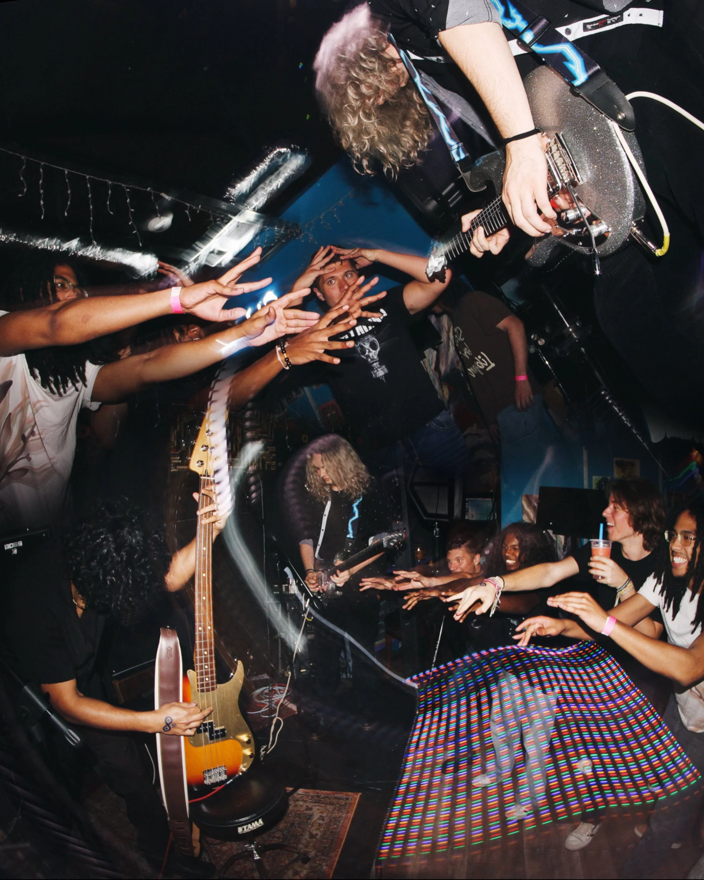 A musician with curly hair playing an electric guitar on stage at a concert, surrounded by enthusiastic audience members reaching out toward the performer, with colorful lighting and a lively atmosphere.