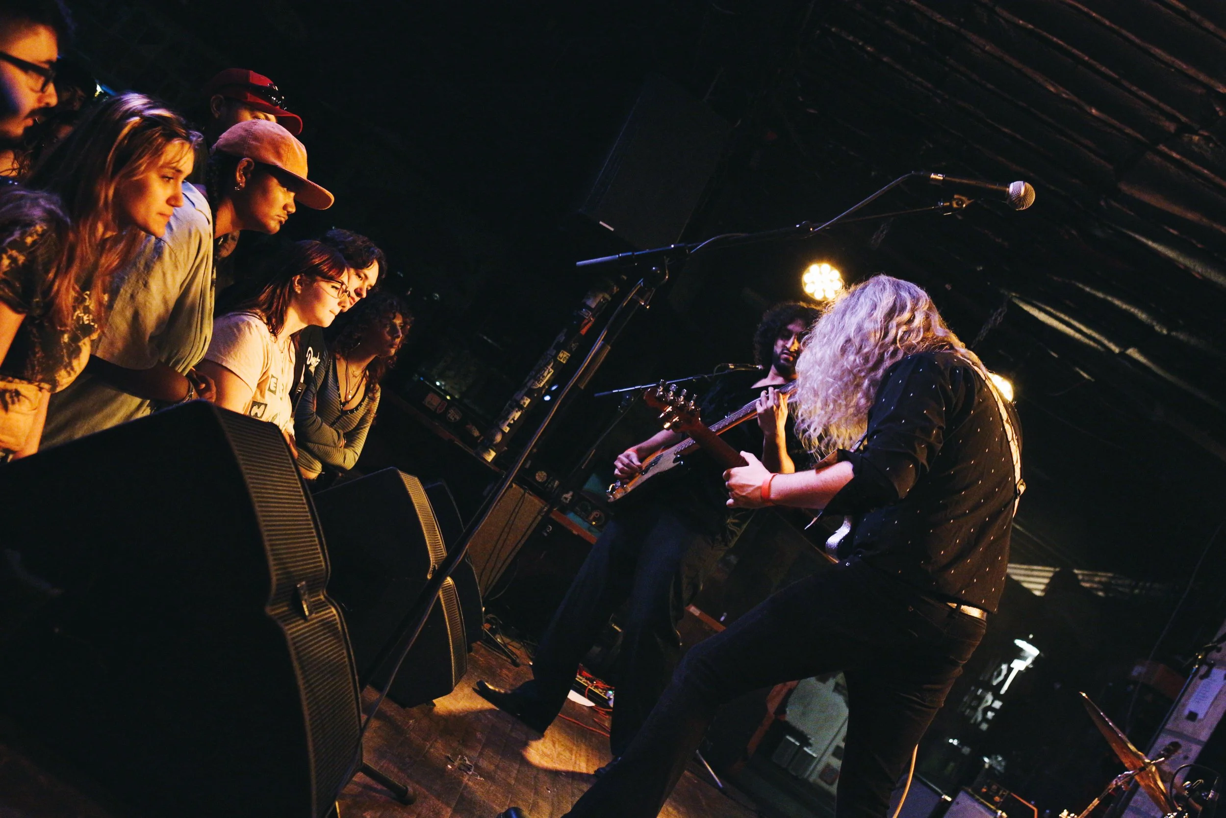 A live music performance with two musicians playing guitars on stage and a group of young people watching attentively.