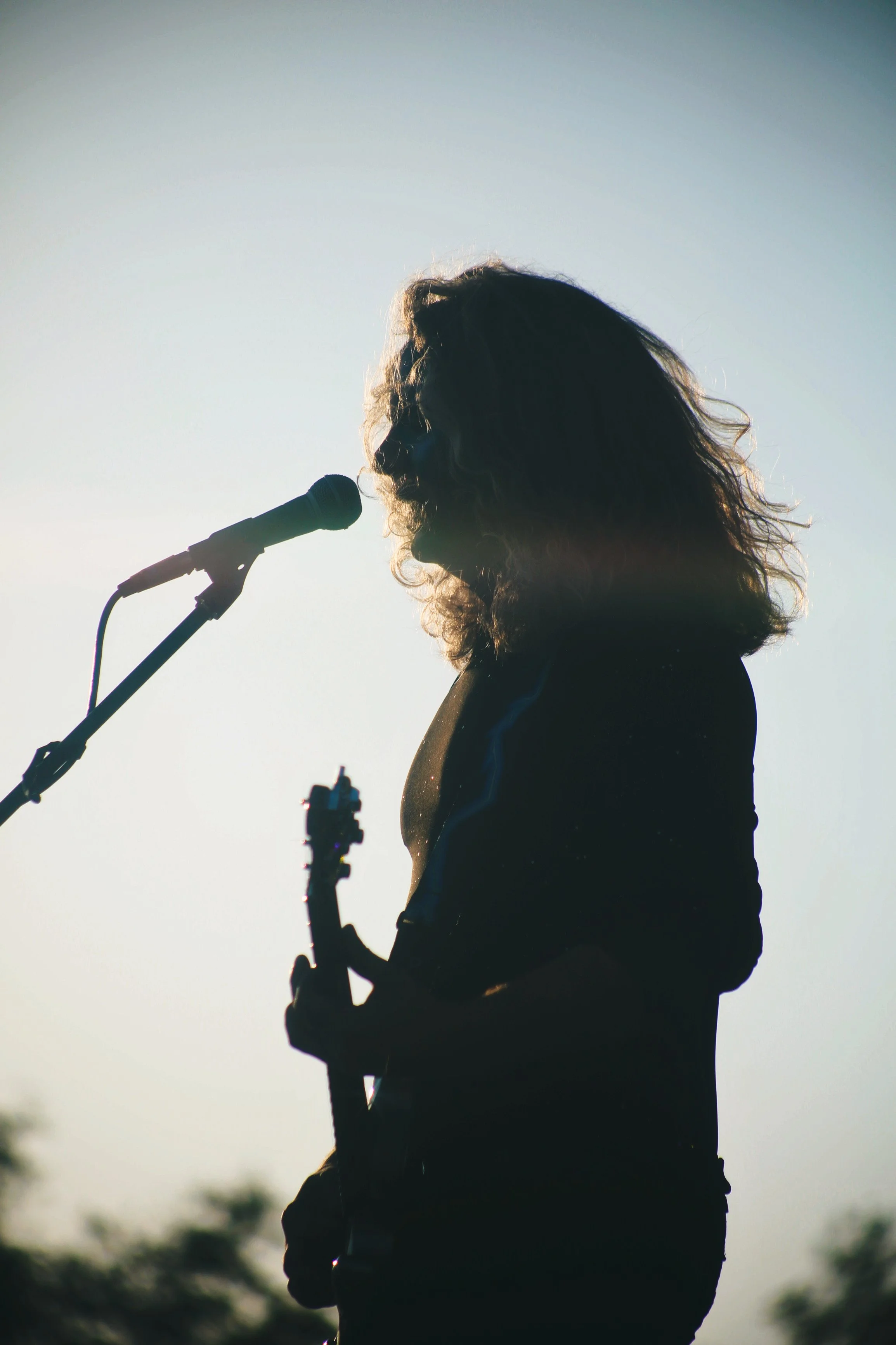 A silhouette of a woman with curly hair singing into a microphone while playing an electric guitar outdoors in front of a bright sky.