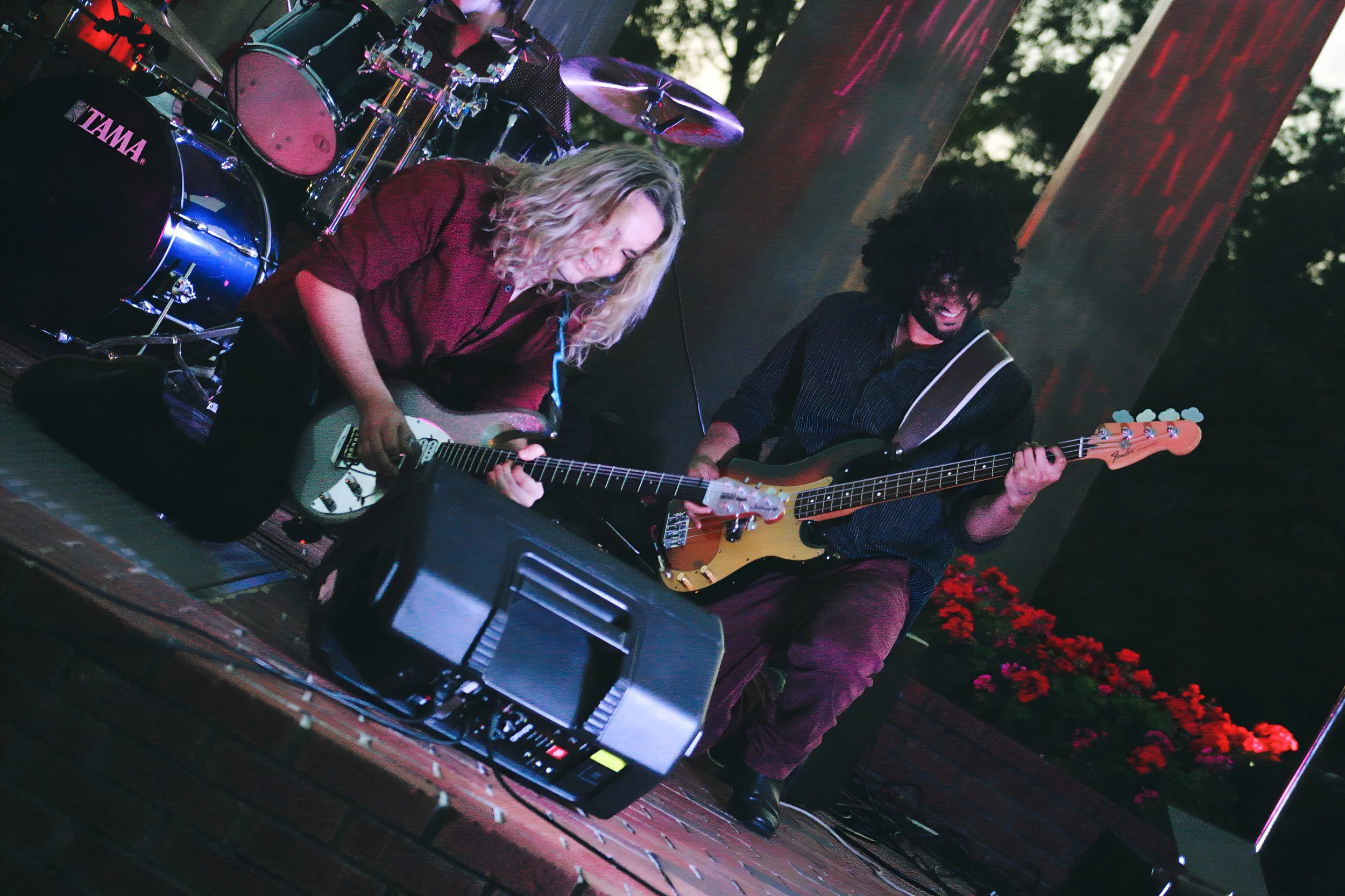 Two musicians, one playing an acoustic guitar and the other playing an electric bass guitar, on a stage with a drum set in the background and red flowers in the distance.