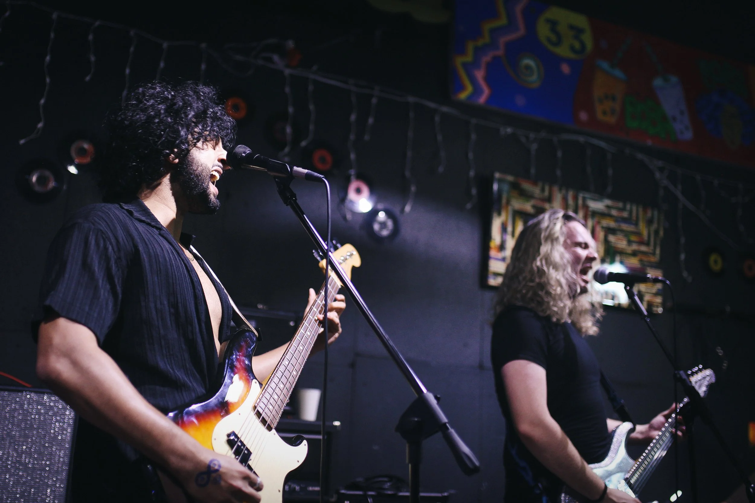 Two musicians performing on stage with guitars and microphones, singing passionately with a dark background and colorful artwork on the wall.