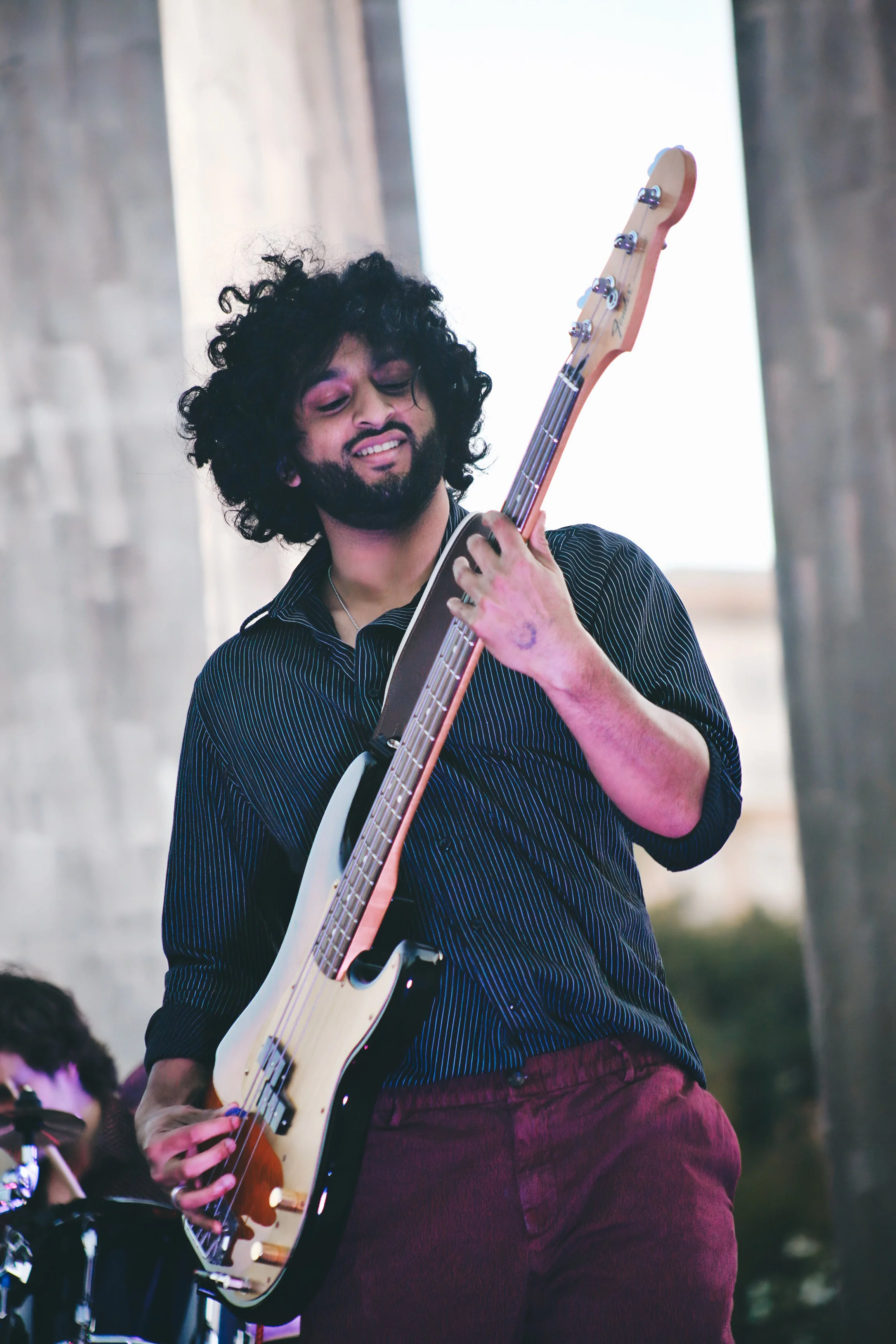 A man with curly black hair and beard playing a bass guitar during a performance, wearing a dark striped shirt and burgundy pants.