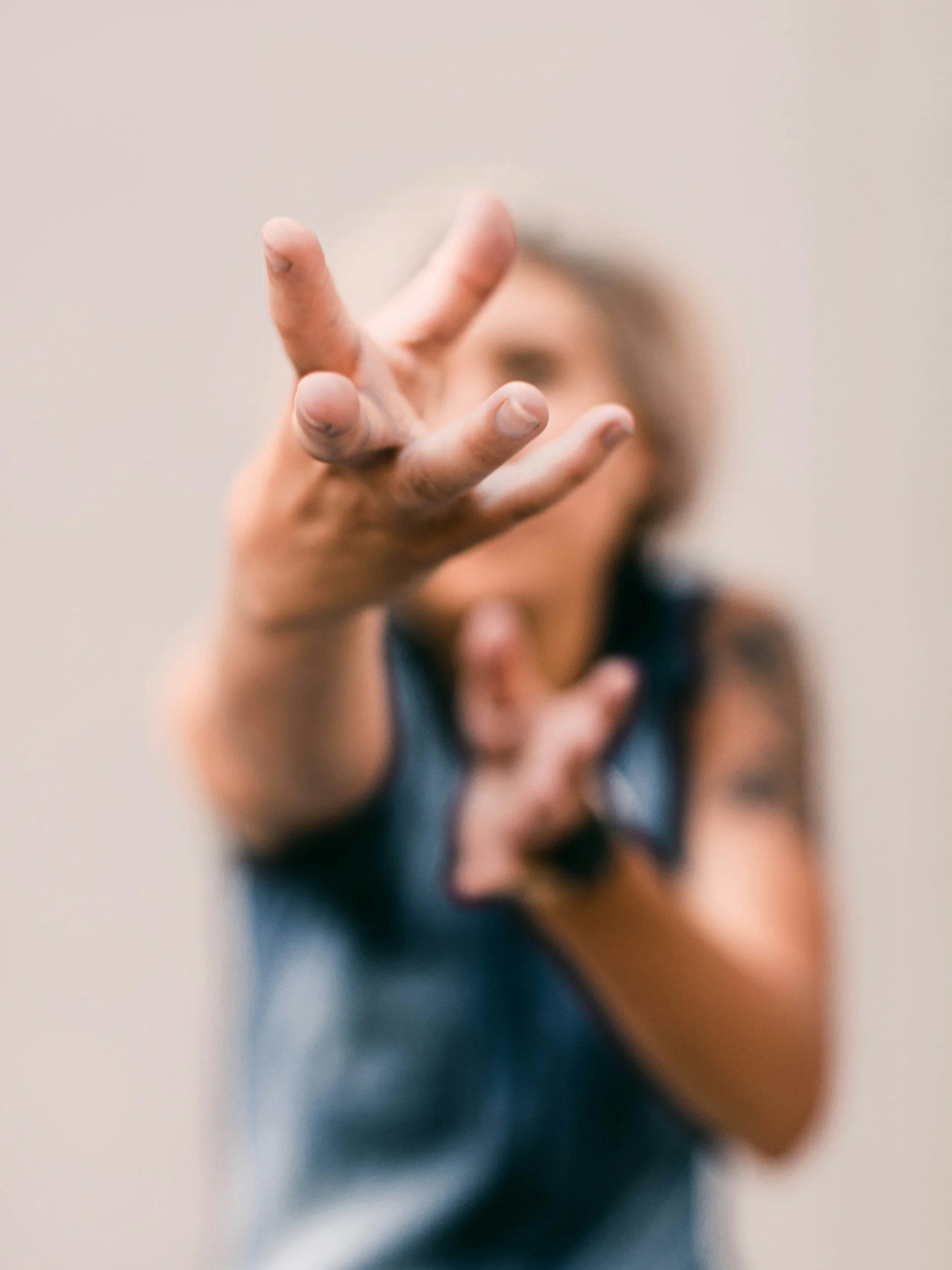 A person with long hair extending their arm toward the camera, making a peace sign with their fingers. The background is plain and blurred.