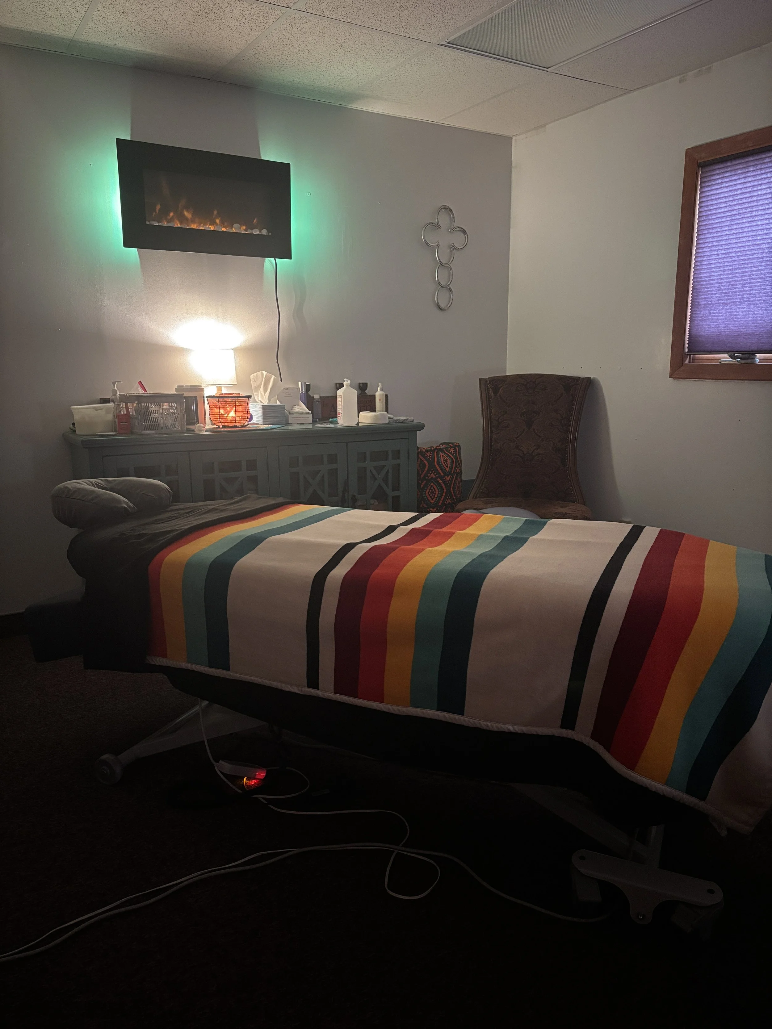 A hospital or medical room with a bed covered in a rainbow-striped blanket, a wheelchair, a wall-mounted electric fireplace, and a wooden framed window with a purple blind. There are various medical supplies and a lamp on a dresser, and a decorative metal wall piece.