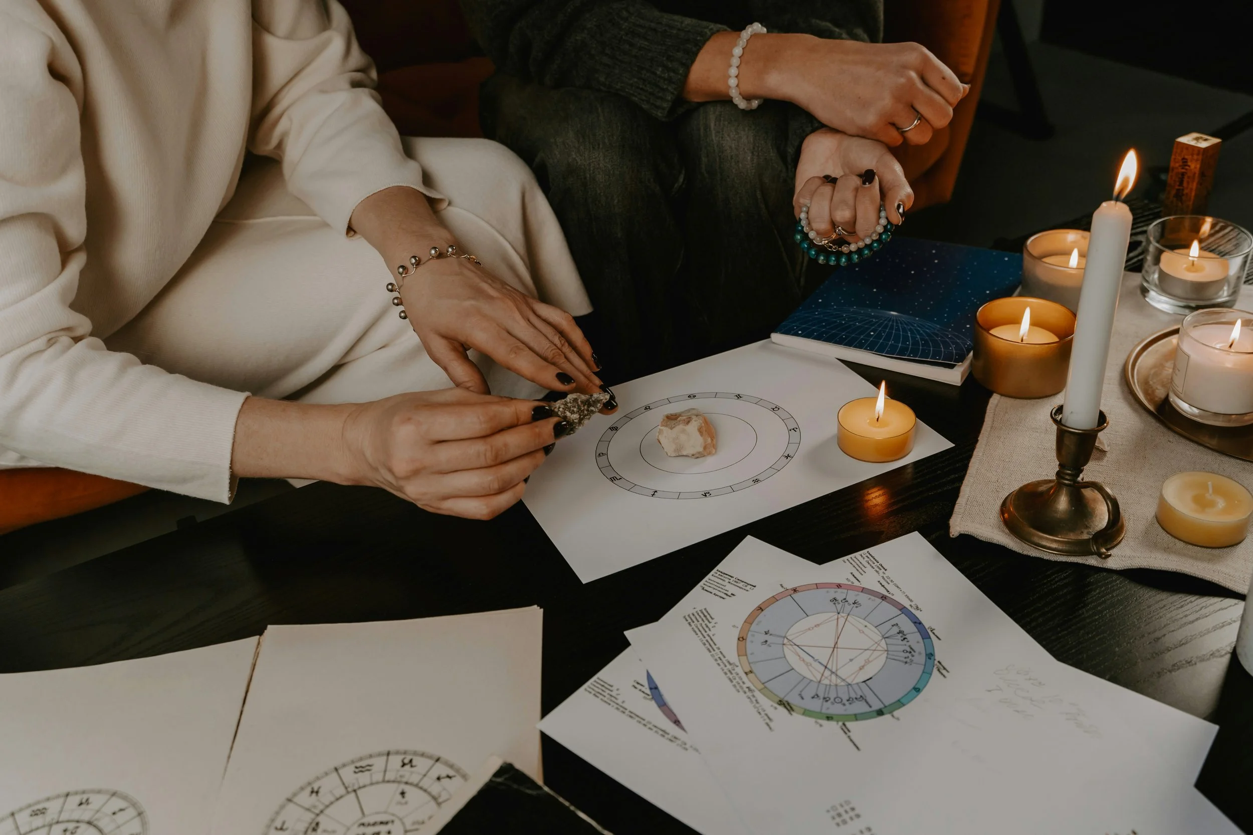 Two people sitting at a table engaged in astrology or spiritual practices. One person wears a cream-colored sweatshirt, the other a dark sweater, and they are holding beaded bracelets. The table is decorated with lit candles, candles in small pots, and astrology charts and notes.