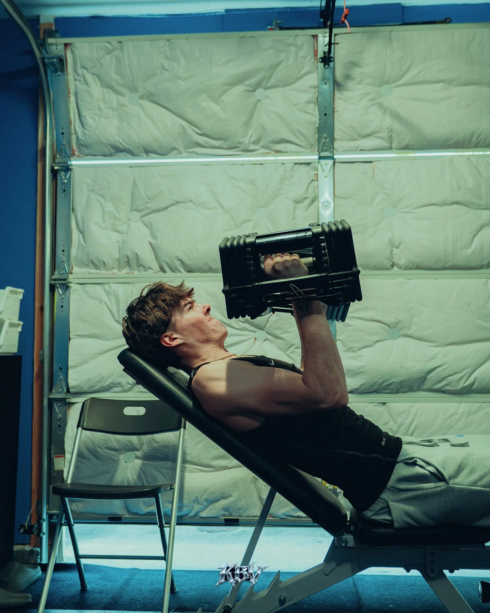 Man performing a chest press with dumbbells on a workout bench in a garage gym.