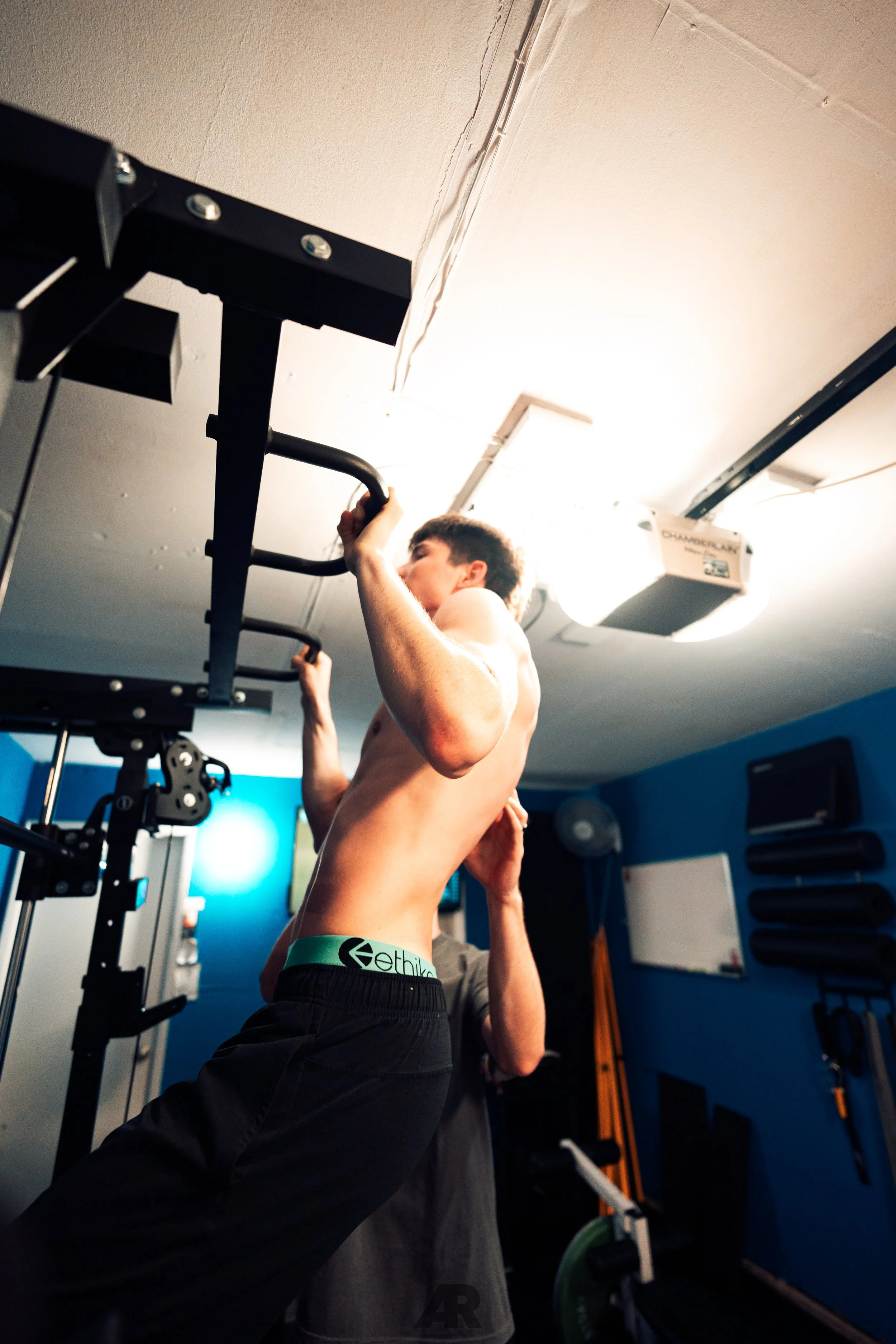 A shirtless man with dark hair doing pull-ups on a black gym pull-up bar in a gym with blue walls. Another person is seen behind him, assisting or spotting.