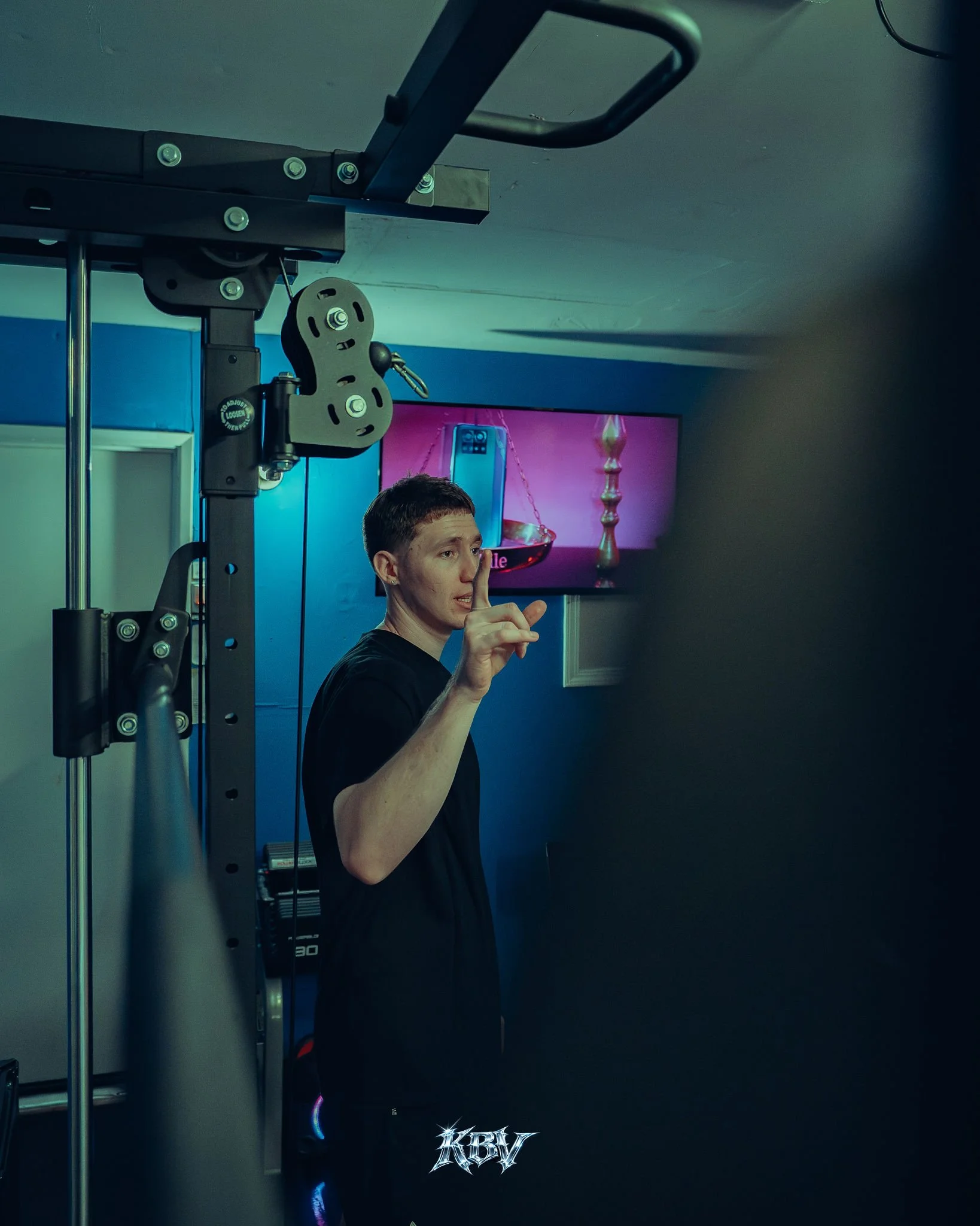 A man with short hair standing near gym equipment, holding up one finger, with a pink screen in the background.