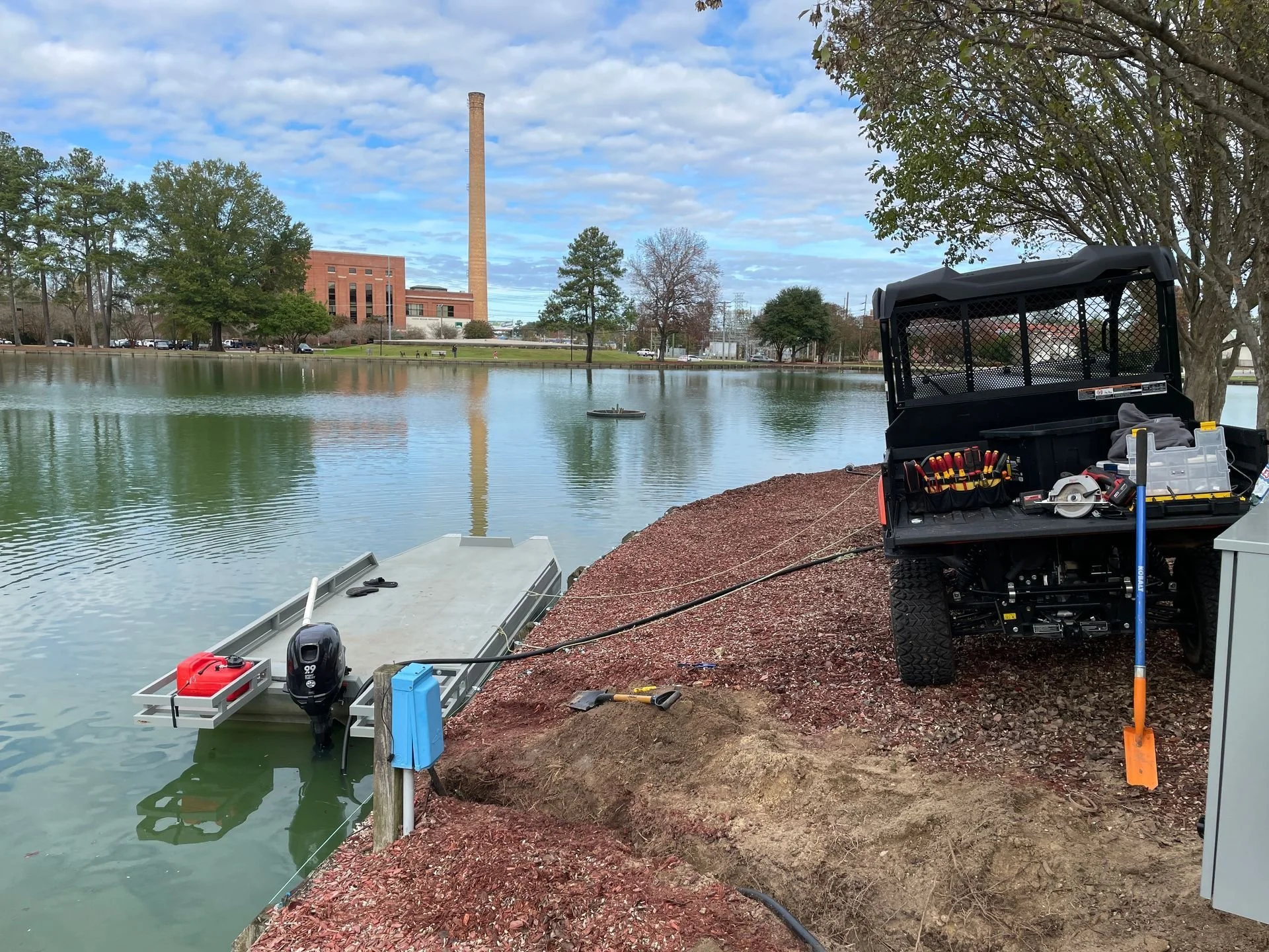 A small boat with a motor attached is docked at the edge of a pond, with tools and equipment on the shore, next to a utility vehicle with a toolbox, on a partly cloudy day.