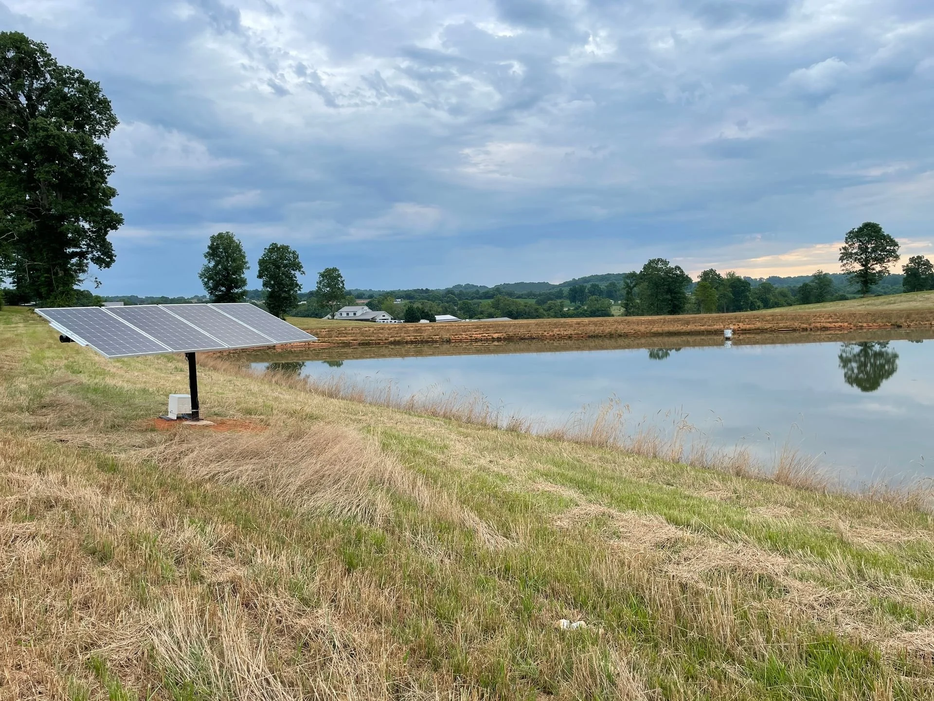 Solar panel installed near a pond in a rural area with trees and cloudy sky in the background.