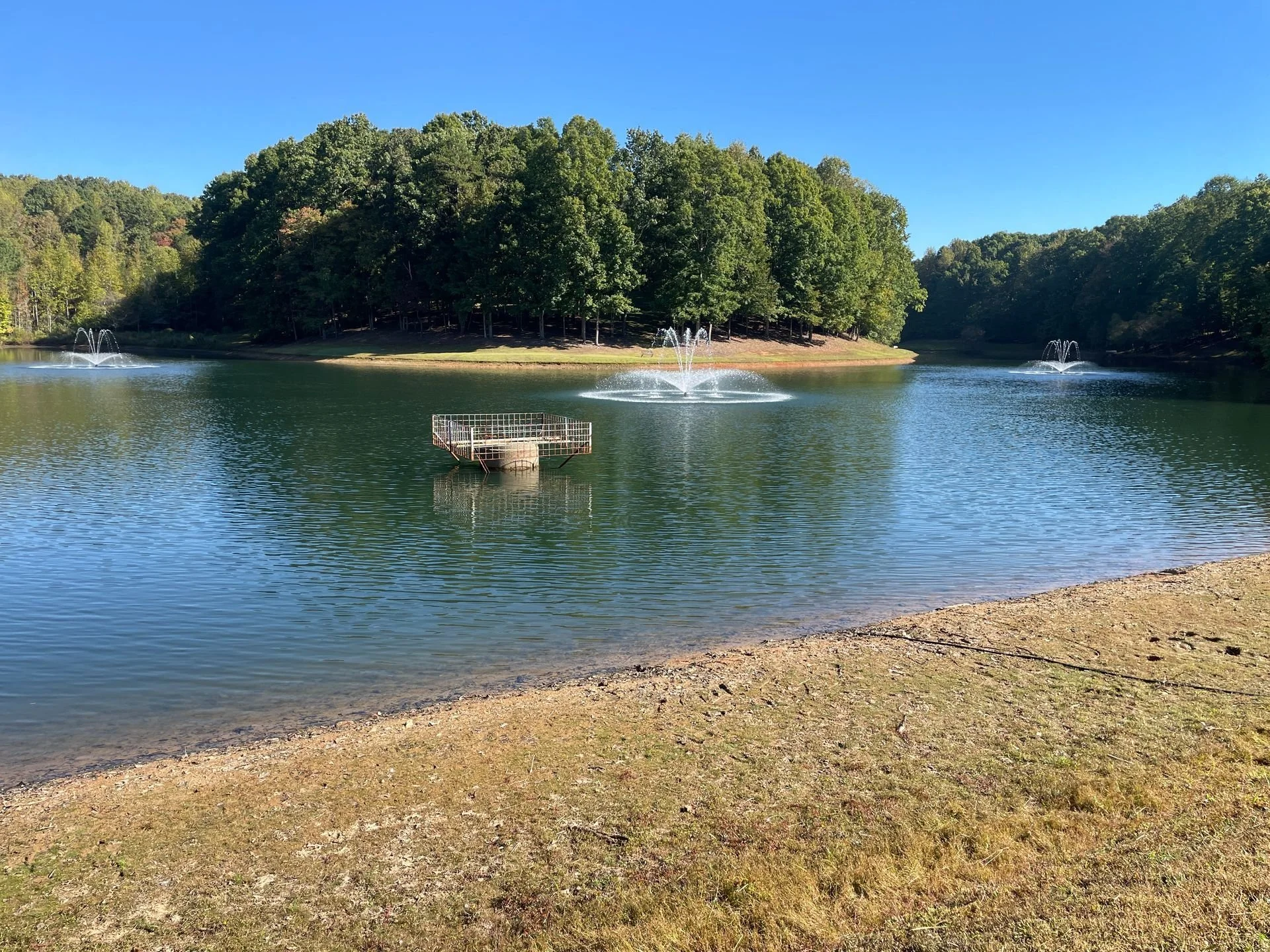 A serene lake surrounded by green trees, with fountains spraying water, and a metal structure in the water near the shore.