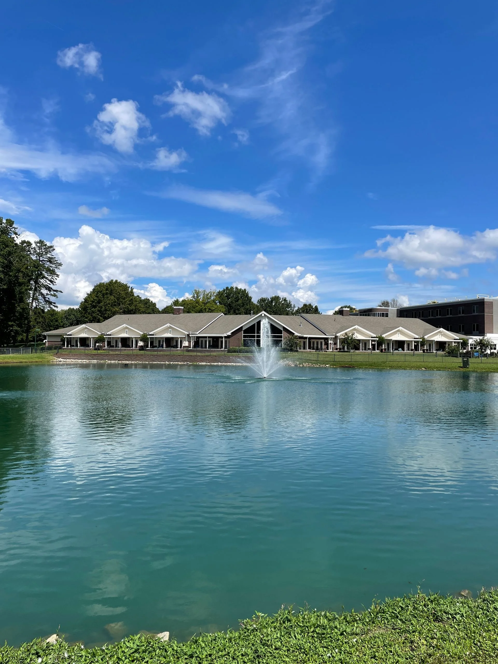 A peaceful pond with a fountain in the center, surrounded by grass, with a building featuring large windows and multiple porches behind it, under a blue sky with clouds.