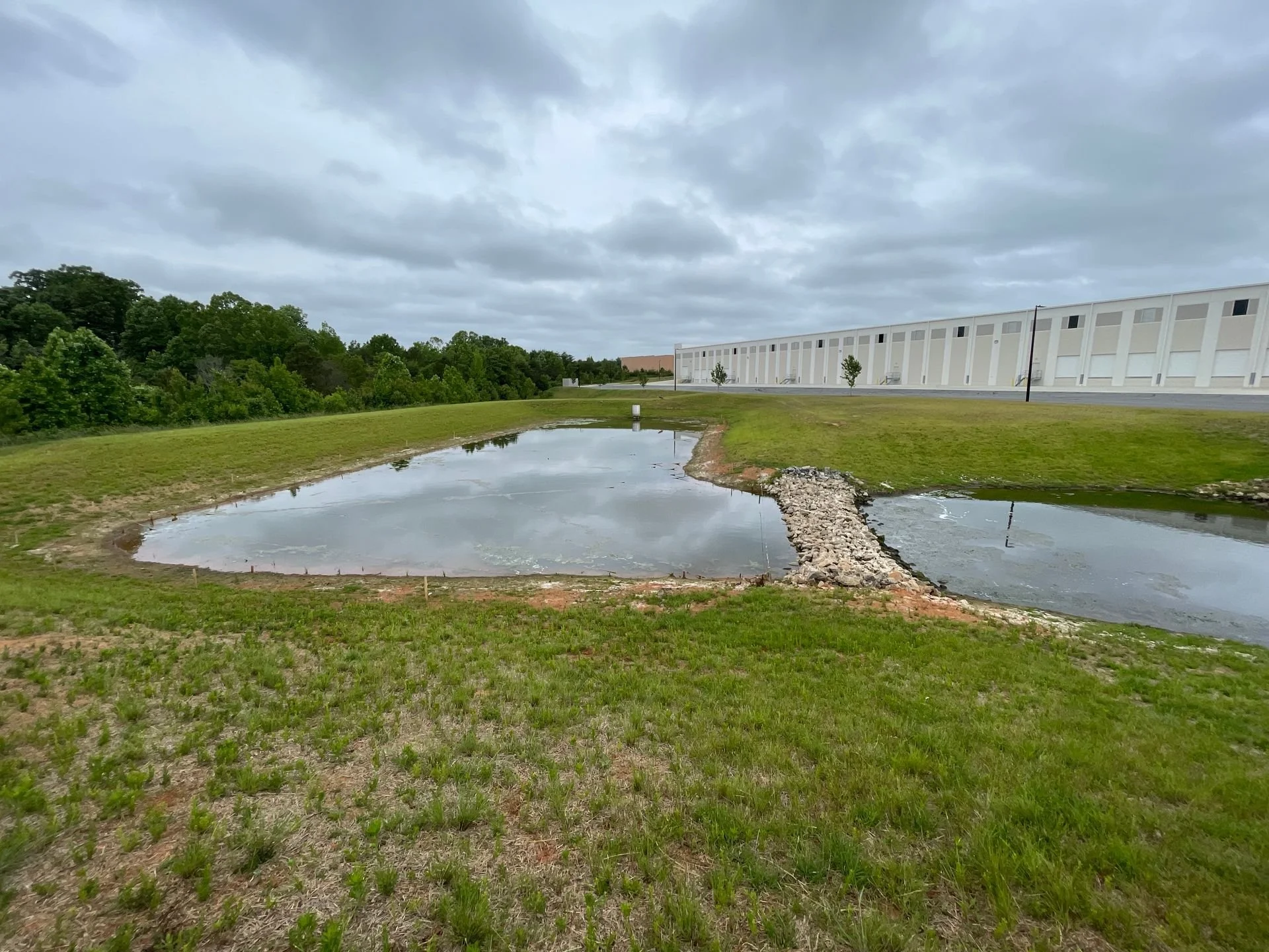 A small pond or water feature with a rocky bridge over it, surrounded by green grass and trees, and an industrial building in the background, under a cloudy sky.