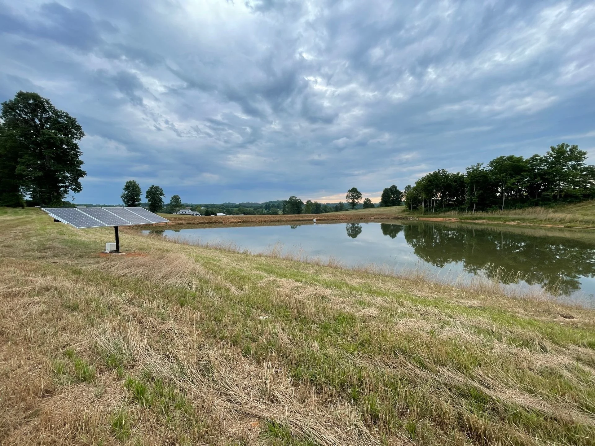 A small pond with calm water reflecting the cloudy sky, surrounded by grassy and wooded areas, with a solar panel installed on the left side.