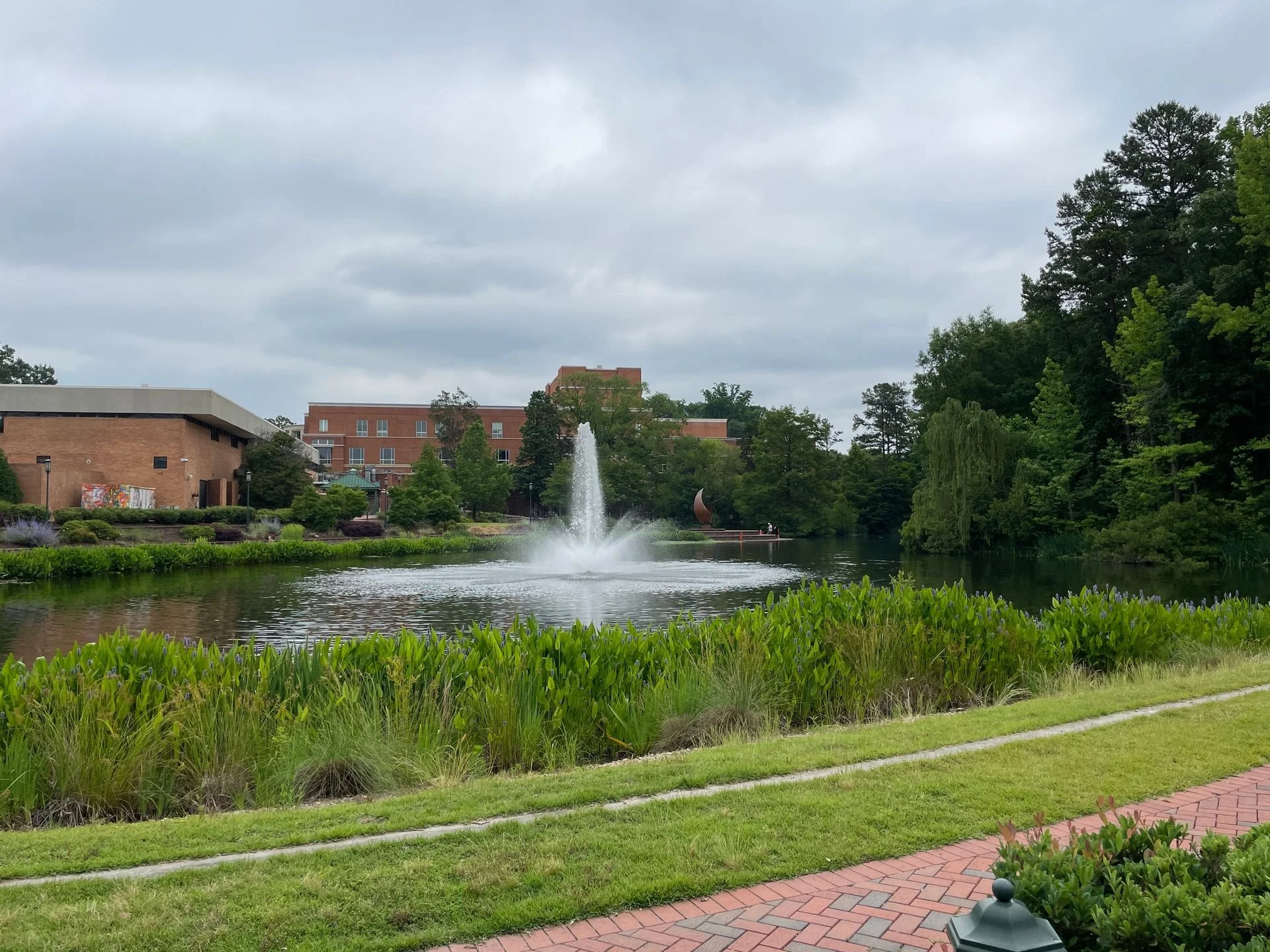 View of a pond with a fountain, surrounded by trees, shrubs, and a brick pathway, with buildings in the background under a cloudy sky.