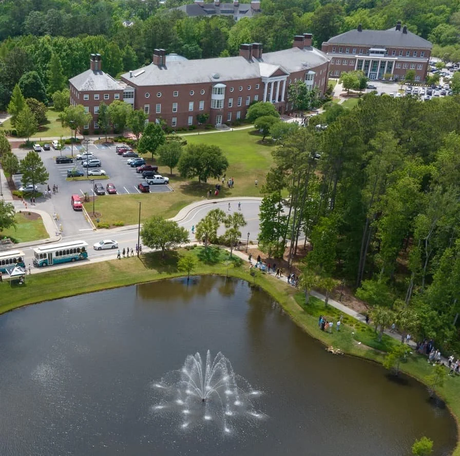 An aerial view of a large brick building surrounded by a parking lot, trees, and a pond with a fountain, along with a group of people walking on the sidewalk.