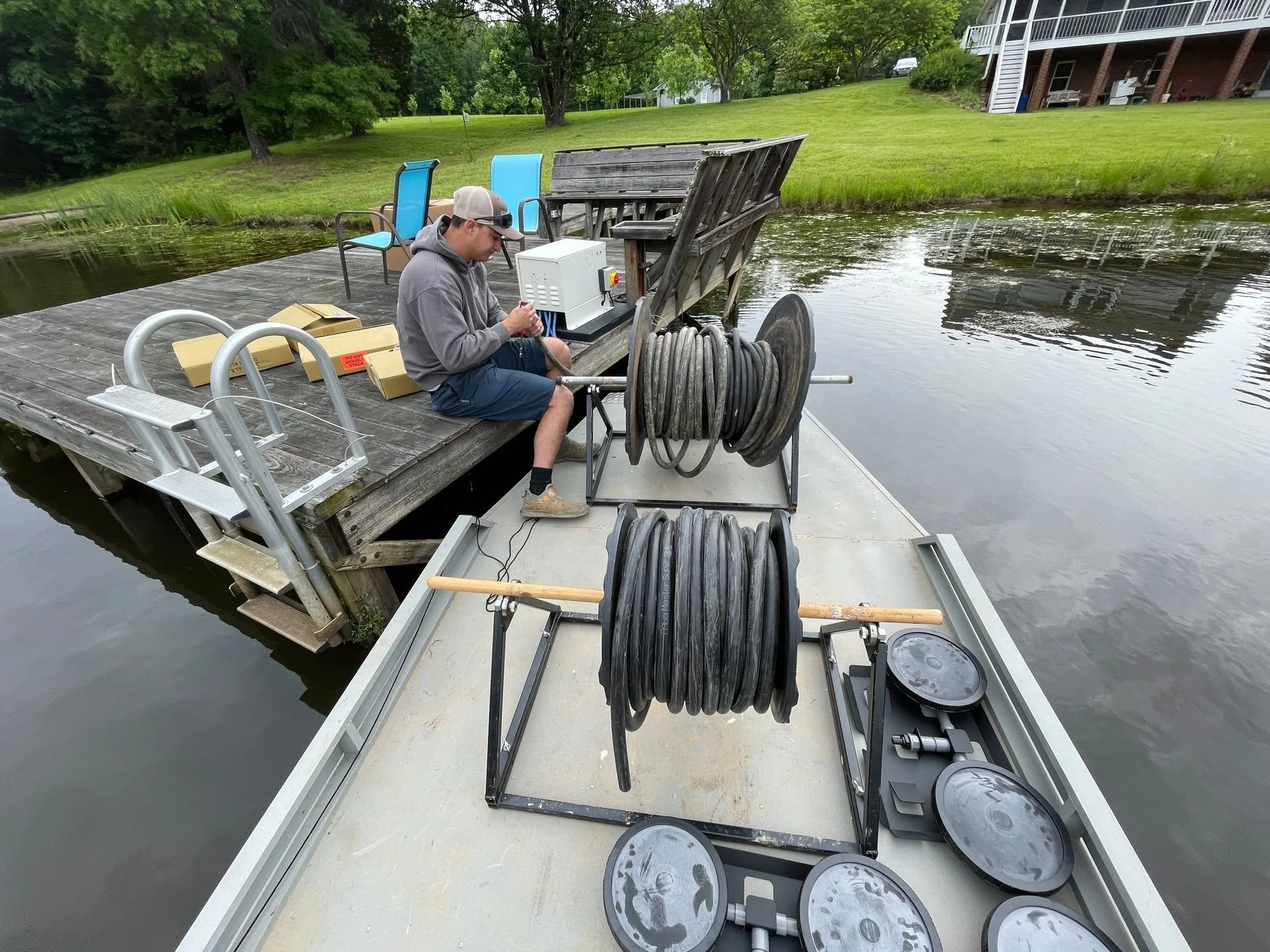 A man sitting on a dock by a lake, wearing a hoodie and shorts, working with electrical or plumbing equipment, with coiled hoses and containers around him, and a house with outdoor seating in the background.