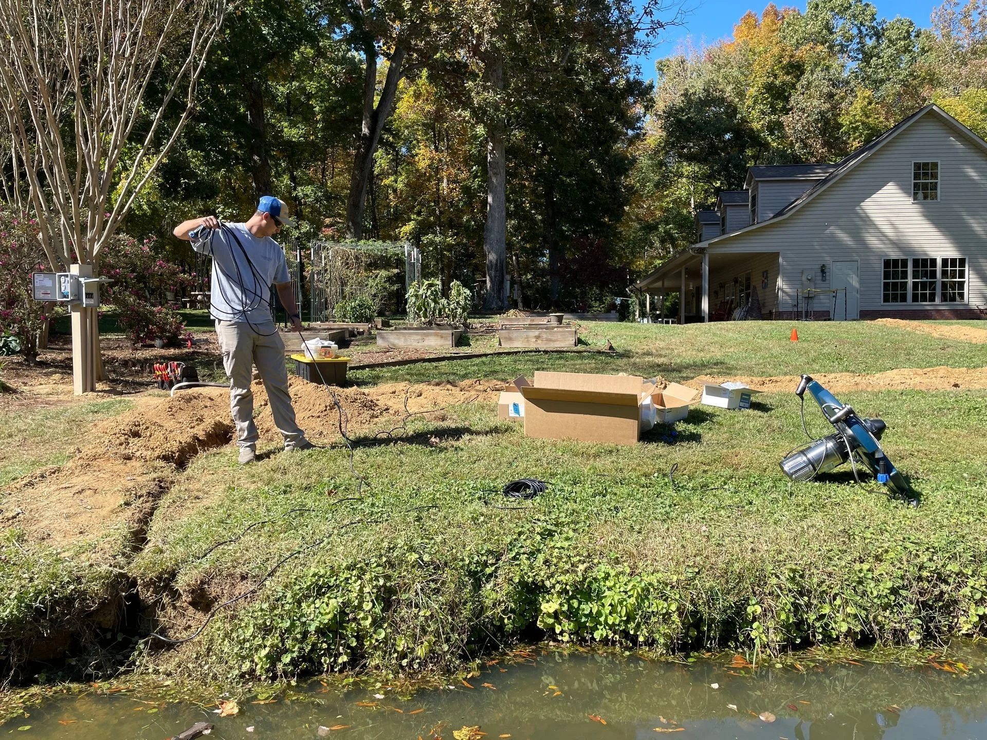 Man working on electrical wiring outdoors near a house with a garden and trees in the background.