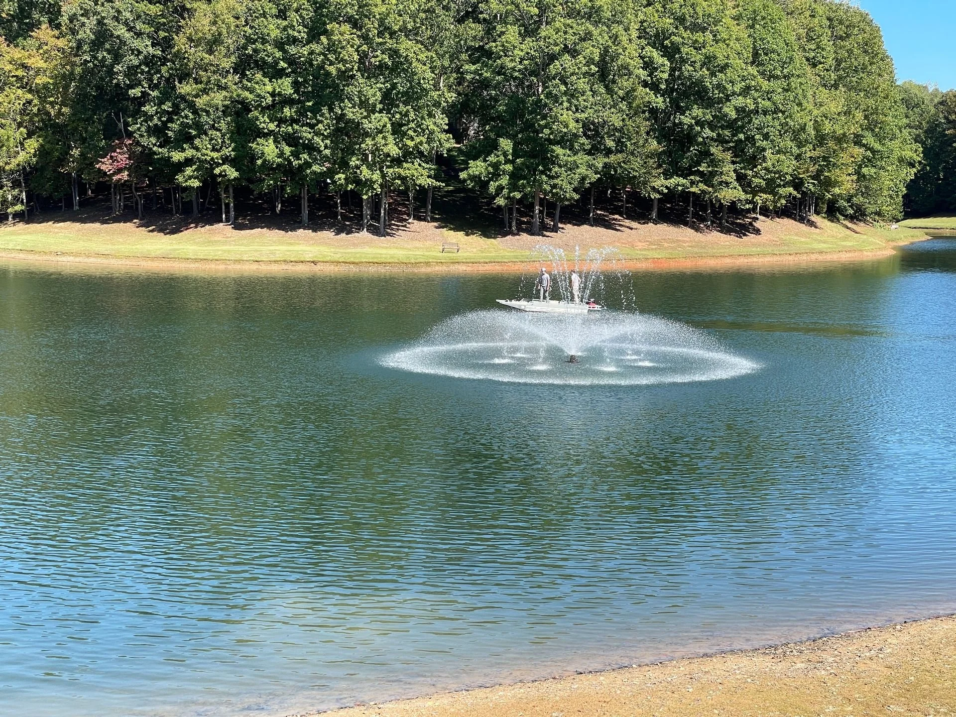 A boat with a person standing on it on a lake, with water being sprayed into the air from the boat's motor. The lake is surrounded by trees and there is a sandy shore in the foreground.