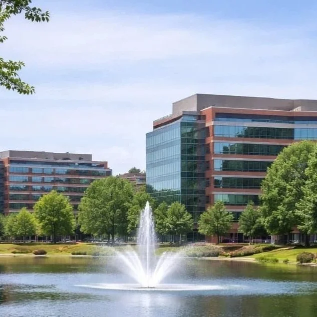 Modern office buildings with reflective glass windows, a fountain in a pond, and green trees in a landscaped area.