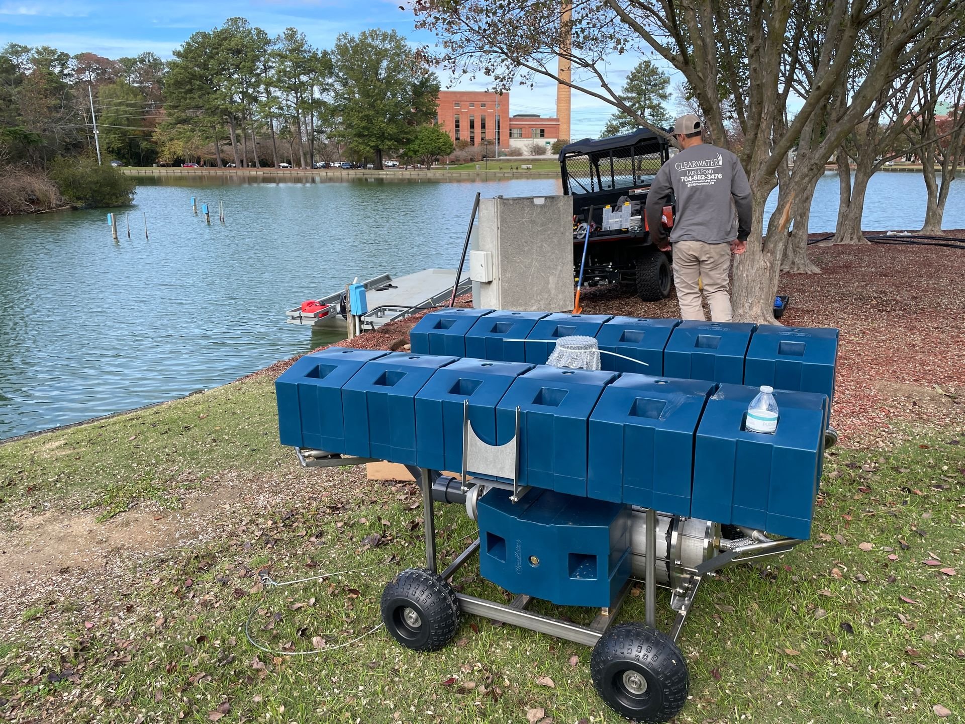 A person standing near a boat dock on a lake, with equipment and containers in the foreground.