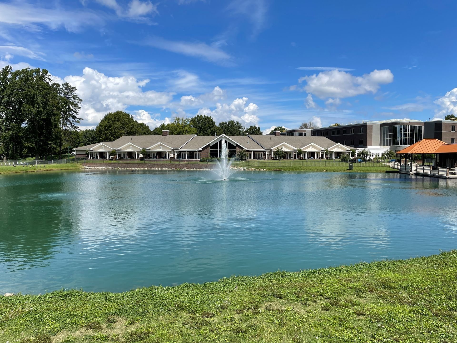 A peaceful pond with a fountain in the center, surrounded by a grassy area and a large building with white columns and a gray roof in the background, under a partly cloudy blue sky.