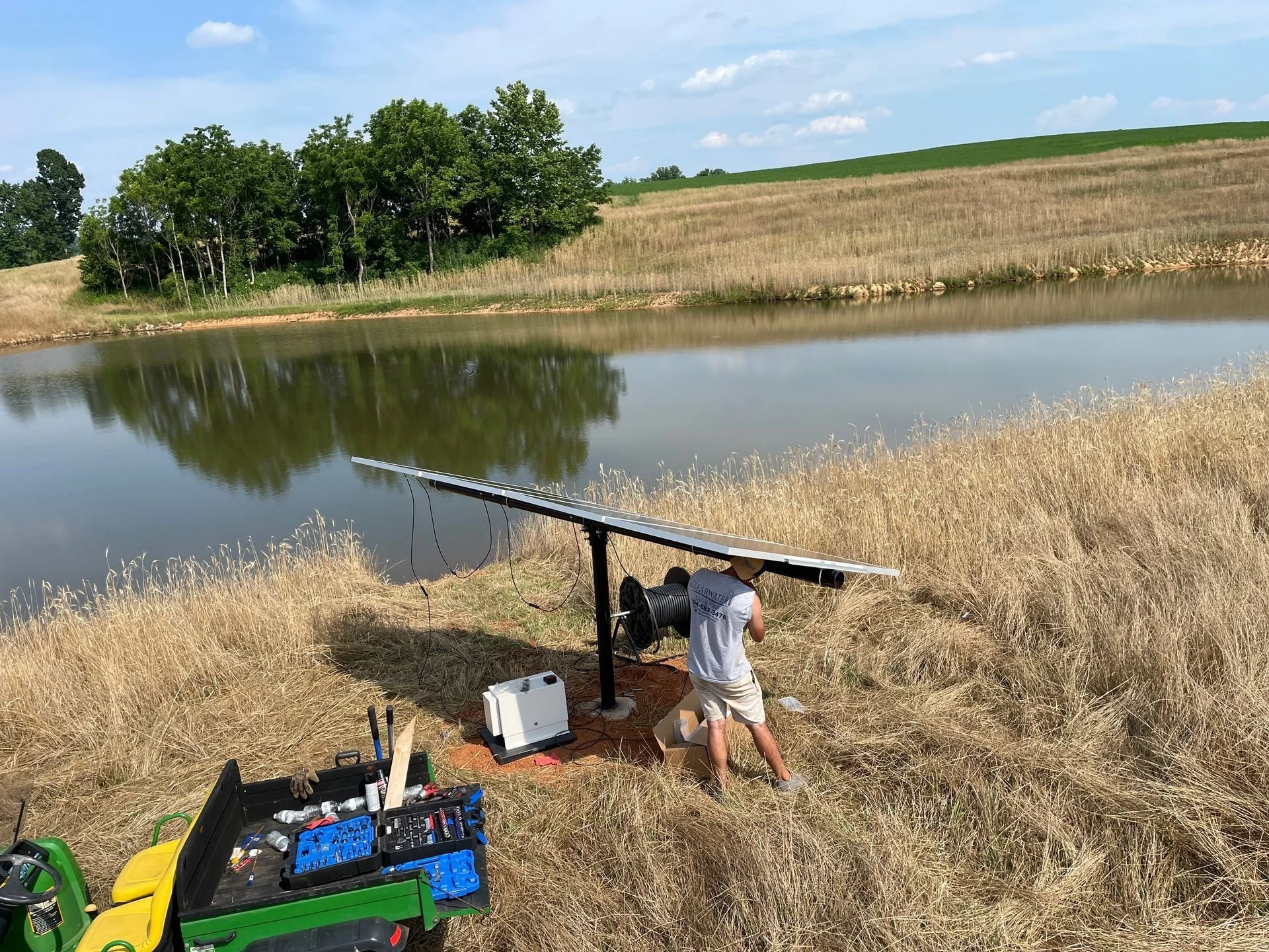 A person working with solar panels near a body of water on a grassy hillside with a toolbox and equipment around, with trees and hills in the background.