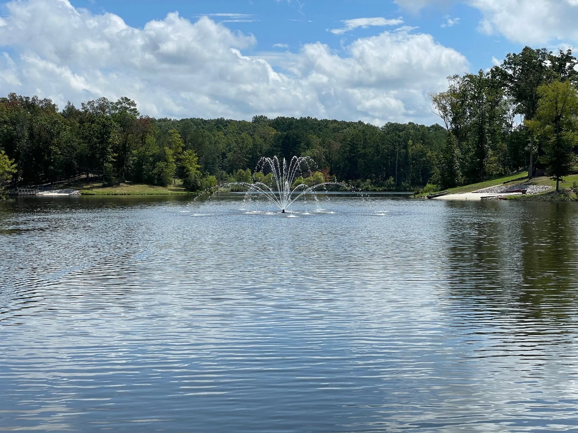 A lake with a fountain in the center, surrounded by green trees and a partly cloudy sky.