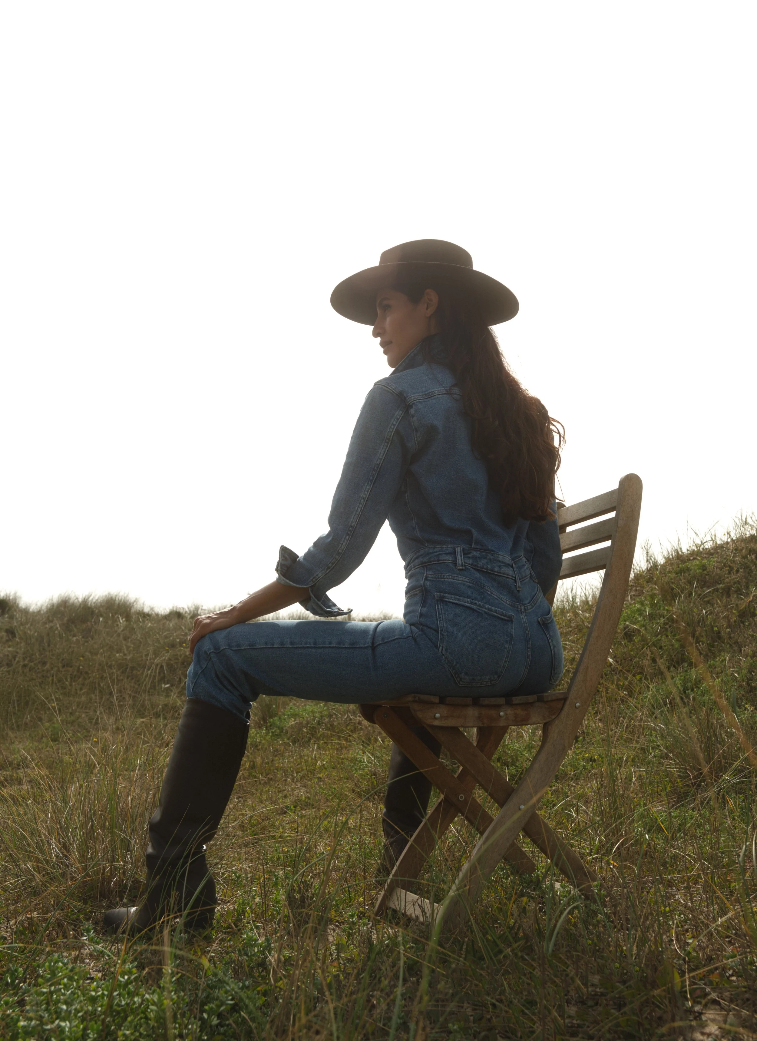 Woman sitting on a wooden chair outdoors in grassy field, wearing a large hat and denim jacket, with long dark hair.