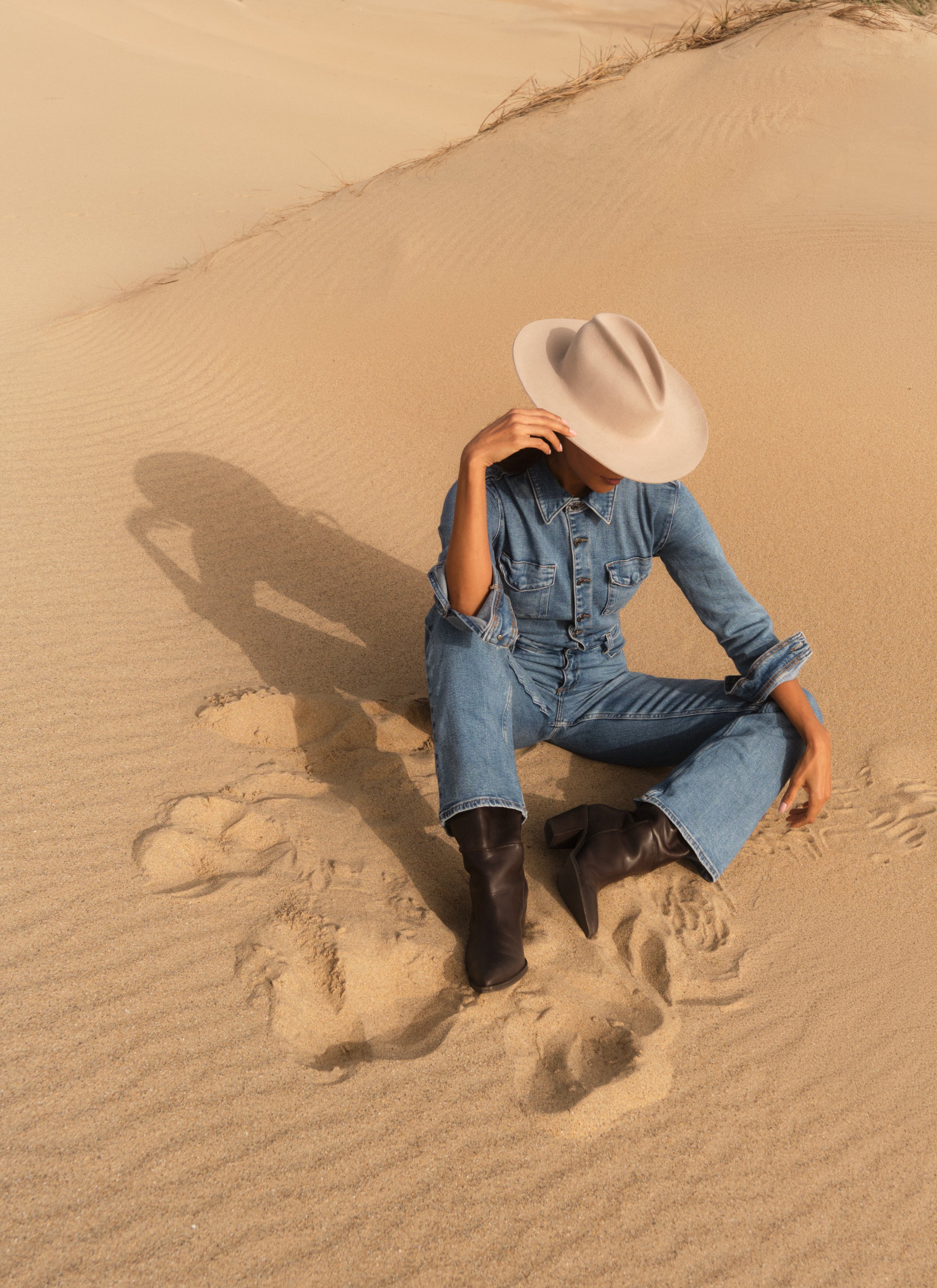 Person sitting on sandy desert ground, wearing denim jeans, a denim jacket, black boots, and a large beige hat, with the shadow cast by the hat visible on the sand.