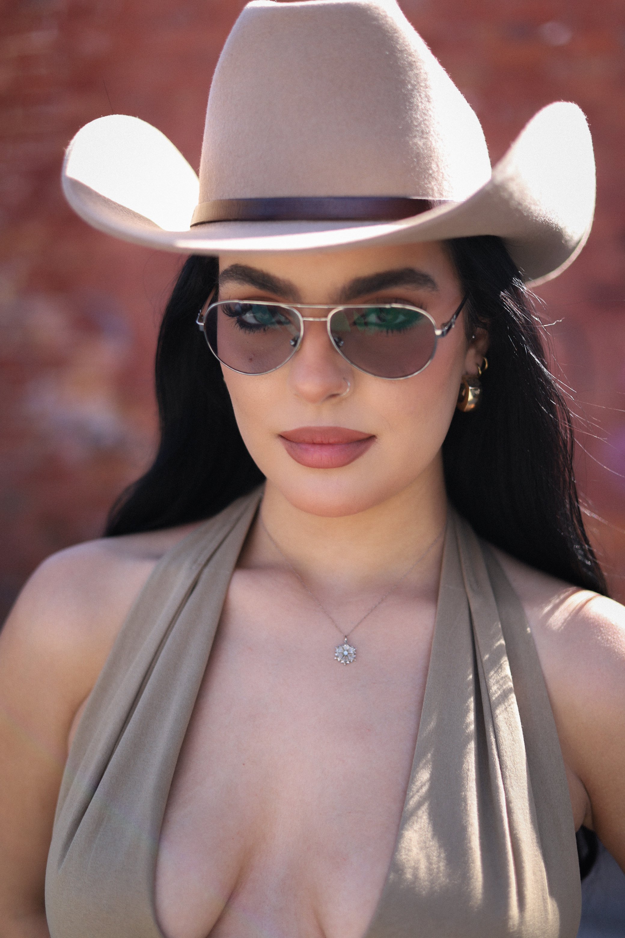 A woman wearing a beige wide-brimmed cowboy hat, sunglasses, and a beige sleeveless top, with dark hair and a necklace, standing outdoors.