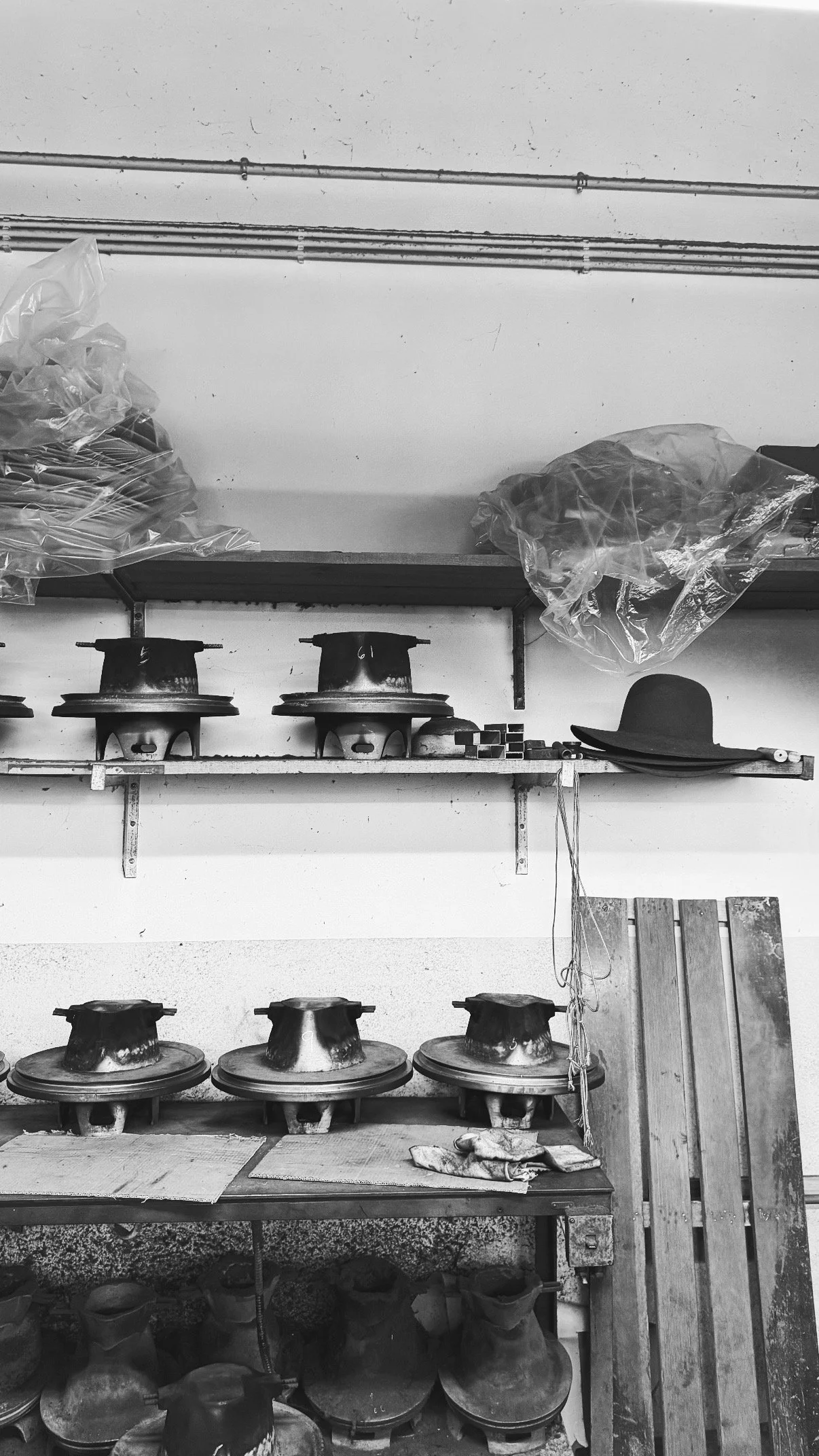A black and white photo showing a workshop with shelves containing hat forms, some wrapped in plastic. There are also hats and materials on the work table, with tools and objects hanging on the wall.