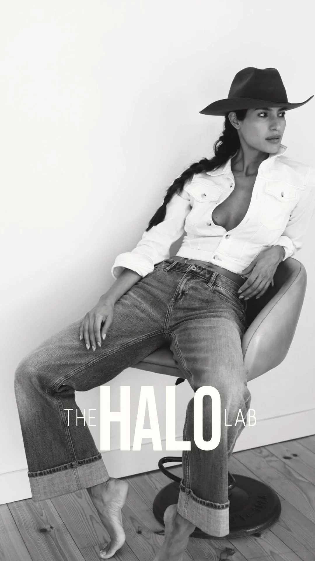 Black and white photo of a woman sitting on a chair, wearing a cowboy hat, white denim jacket, and jeans, with her legs crossed and hand resting on her thigh.