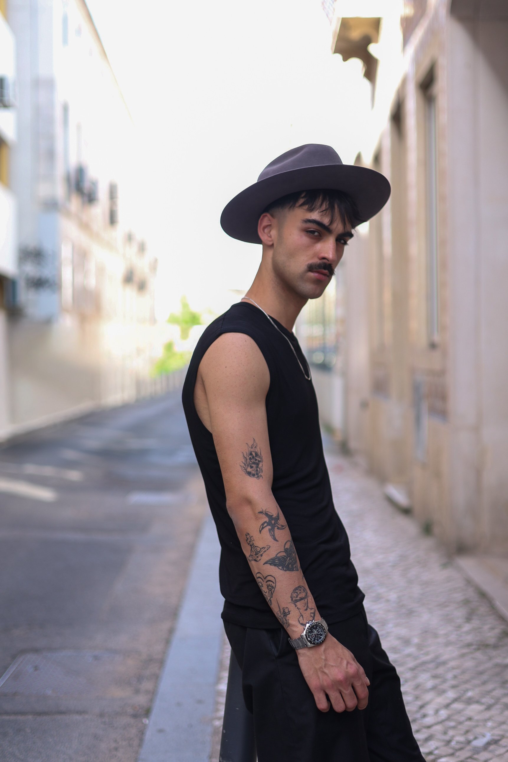 A young man with tattoos on his arms, wearing a black sleeveless shirt, a silver wristwatch, and a wide-brimmed gray hat, stands on a city street. He has dark hair, a mustache, and a confident expression as he looks at the camera. The background features blurred buildings on either side of the street.