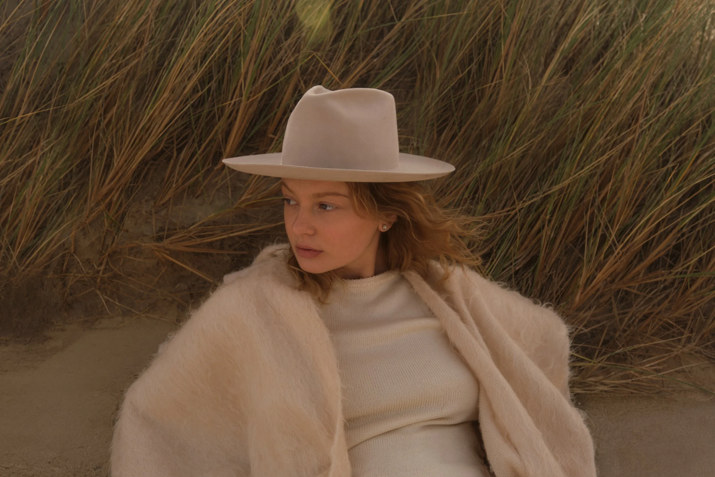 A woman with red hair wearing a wide-brimmed hat, a cream-colored fuzzy coat, and a white top, sitting among tall brown grass.