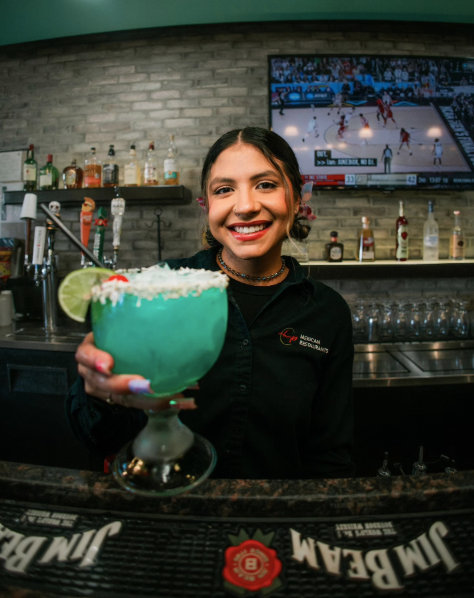 Smiling bartender holding a blue cocktail garnished with lime and coconut, behind a bar with liquor bottles and a TV showing a hockey game.