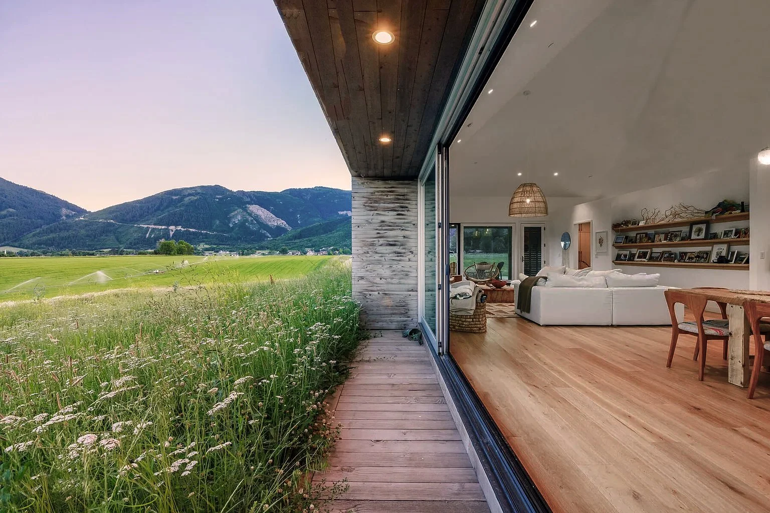 Open-concept living room with white sofa, wooden flooring, and large sliding glass doors opening to a view of green fields and mountains.
