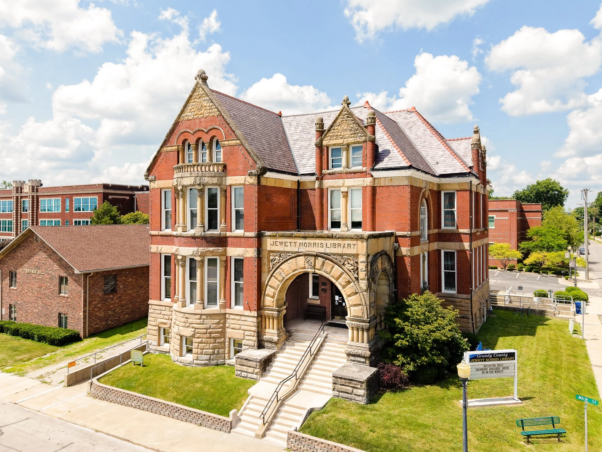 Front view of a historic red brick building with stone accents and an arched entrance labeled "Jewett Norris Library" under a partly cloudy sky. There are stairs leading to the entrance and well-maintained grass and trees around.