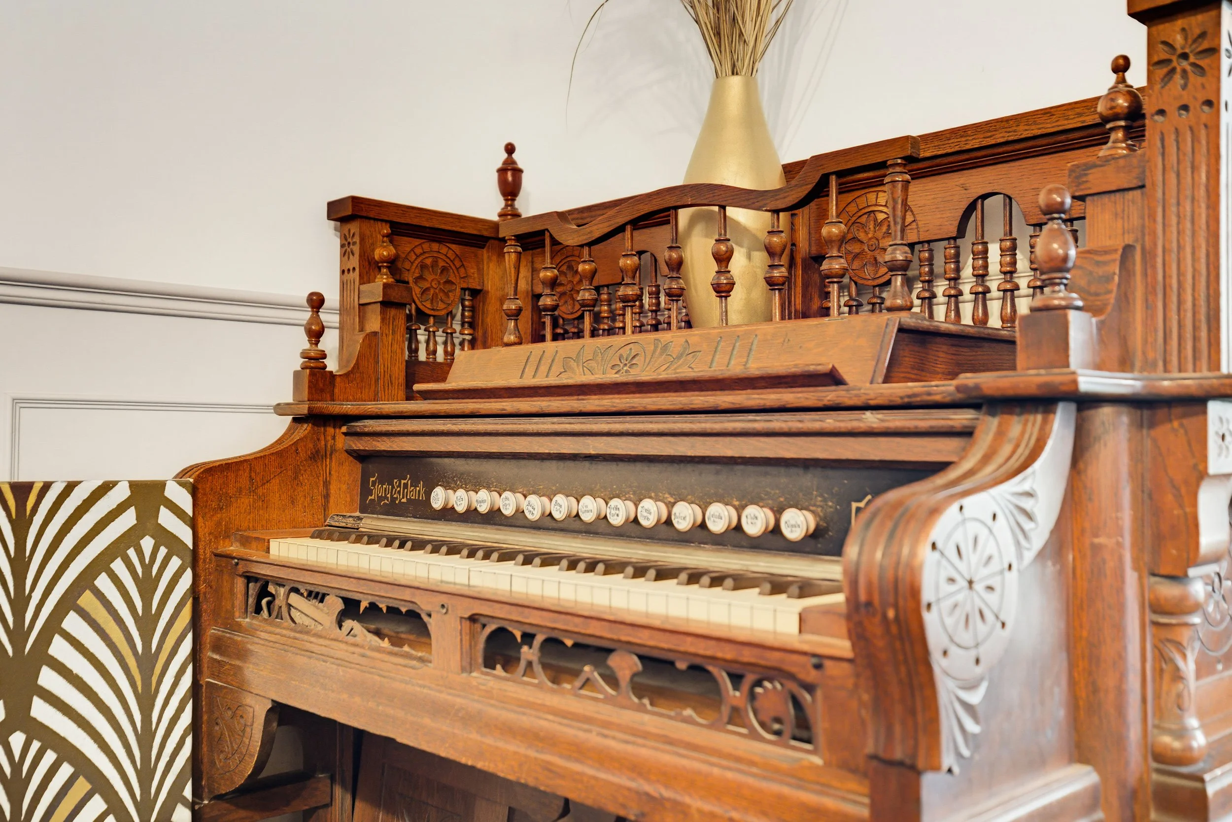 A vintage wooden church pipe organ with decorative carvings, a row of white and black keys, and a large beige vase with dried decorative grasses on top.