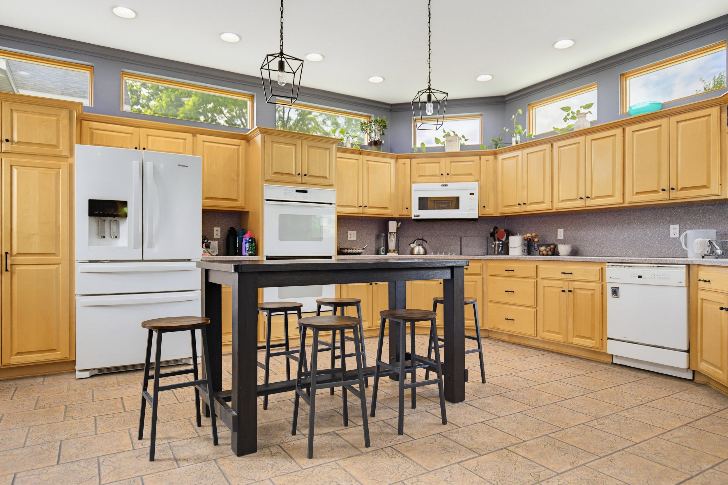 Kitchen with wooden cabinets, white appliances, black island table with stools, and large windows with green trees outside, illuminated by ceiling lights and pendants.