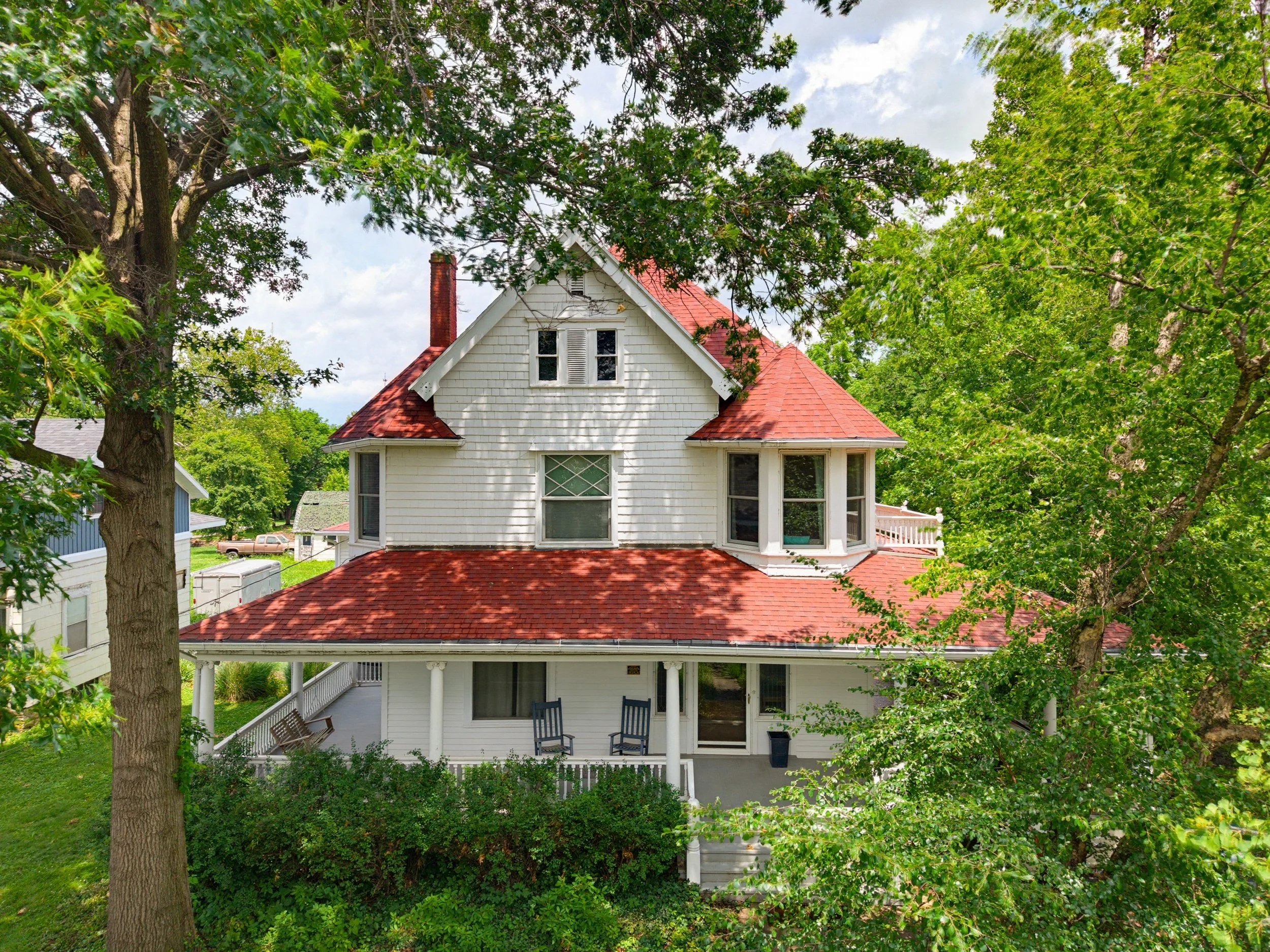 A white two-story house with a red shingled roof and gabled dormer windows, partially obscured by large green leafy trees, with a porch and multiple windows.