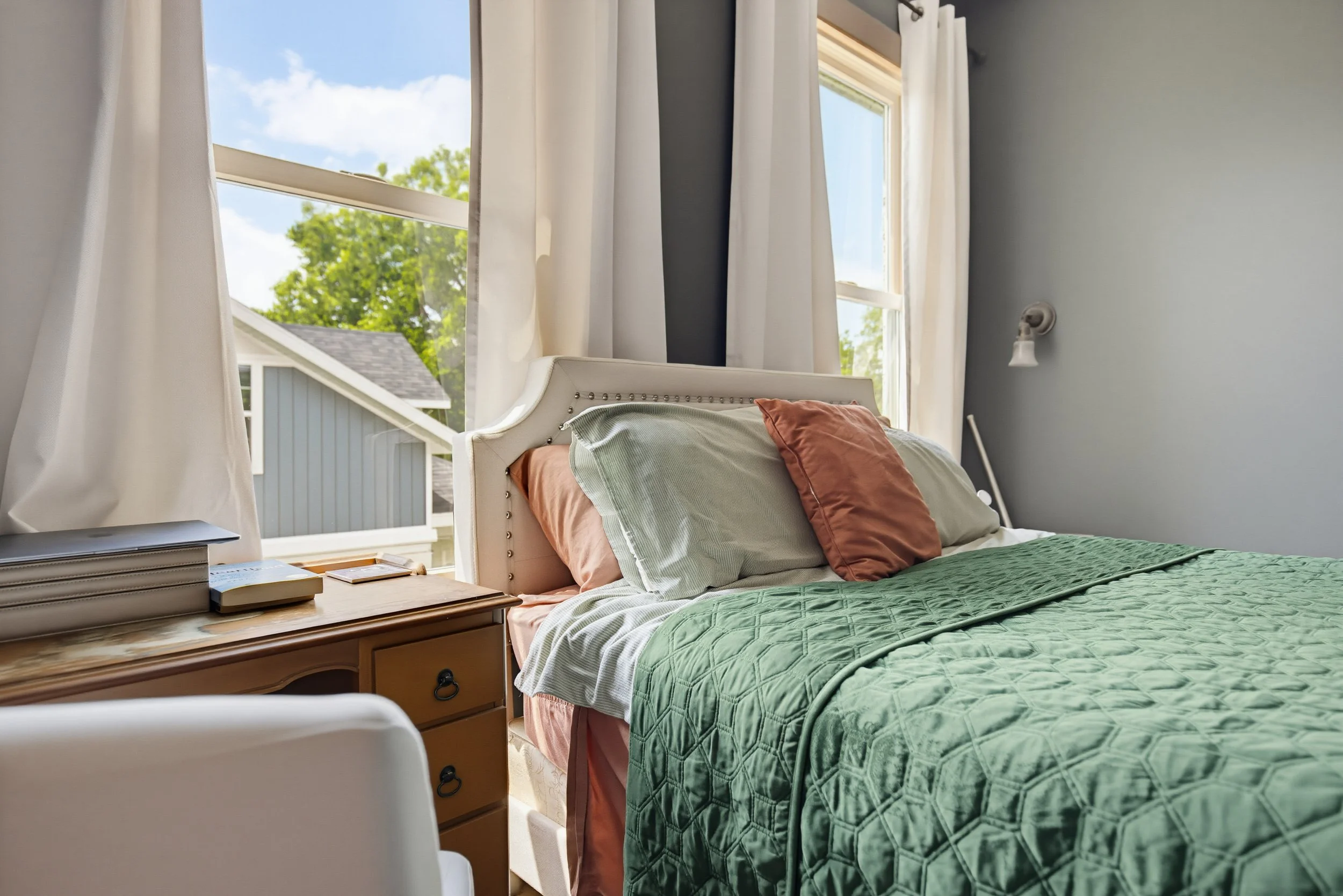 A bedroom with a bed covered in green quilt and pink pillows, beside a wooden nightstand with books, near a large window with white curtains showing a sunny day outside.