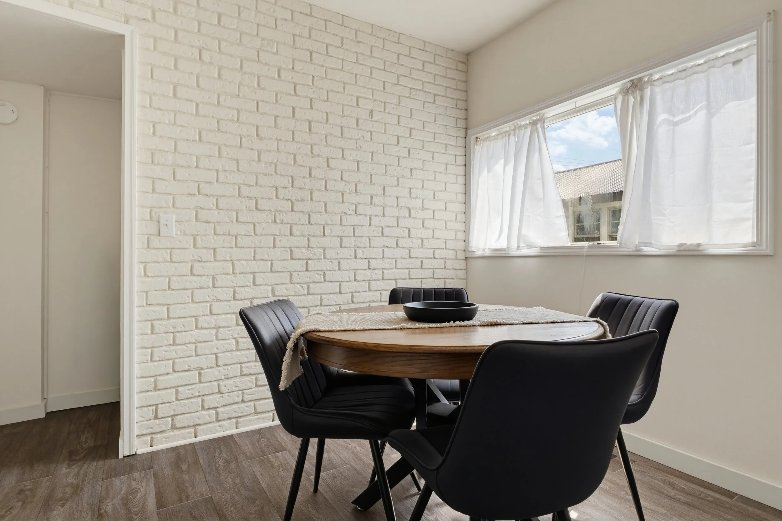 Dining area with a round wooden table, black chairs, a white brick wall, and a window with white curtains showing a sunny sky and nearby rooftops.