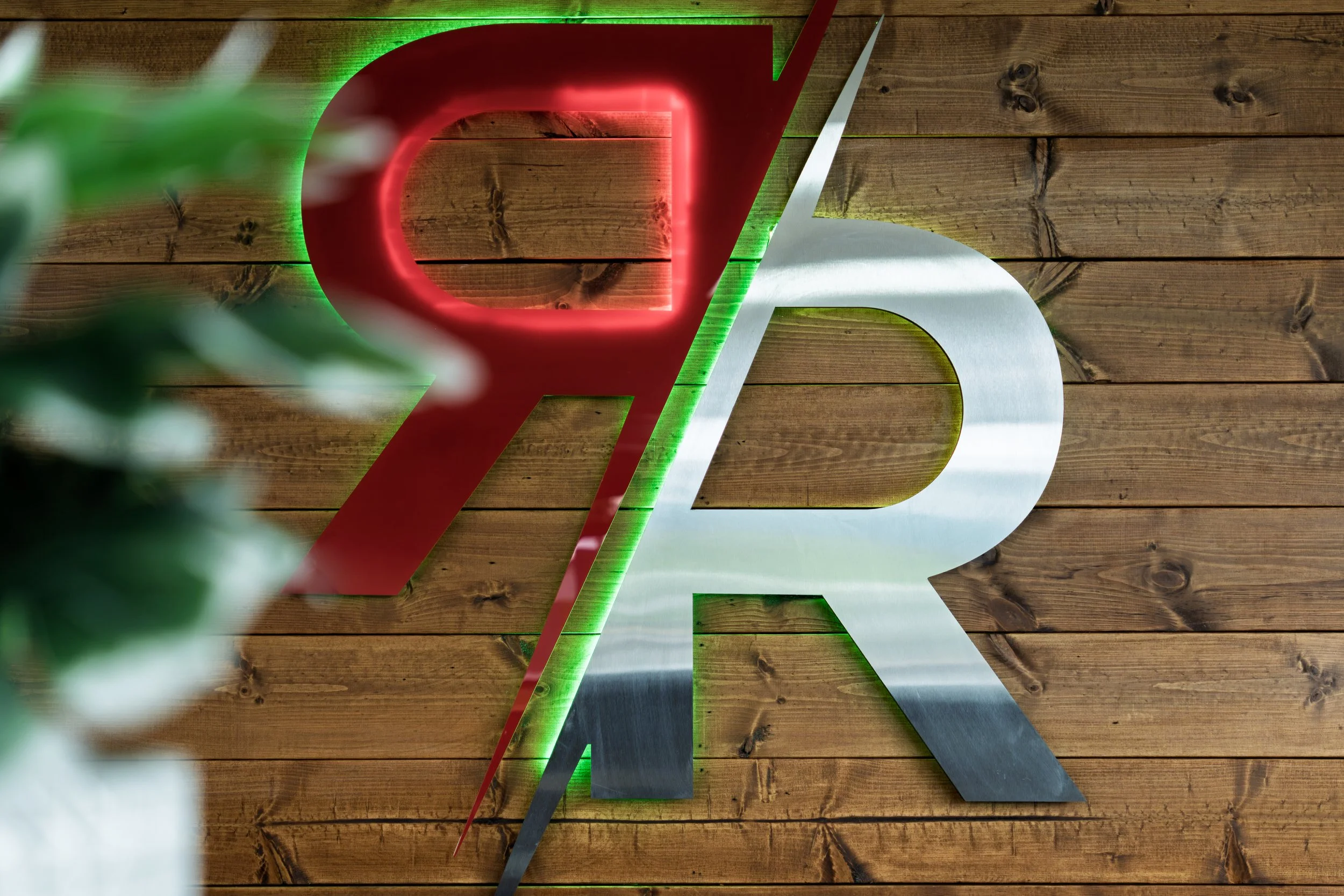 Close-up of a modern geometric hanging sign on a wooden wall, with illuminated edge lighting and blurred green plant leaves in the foreground.