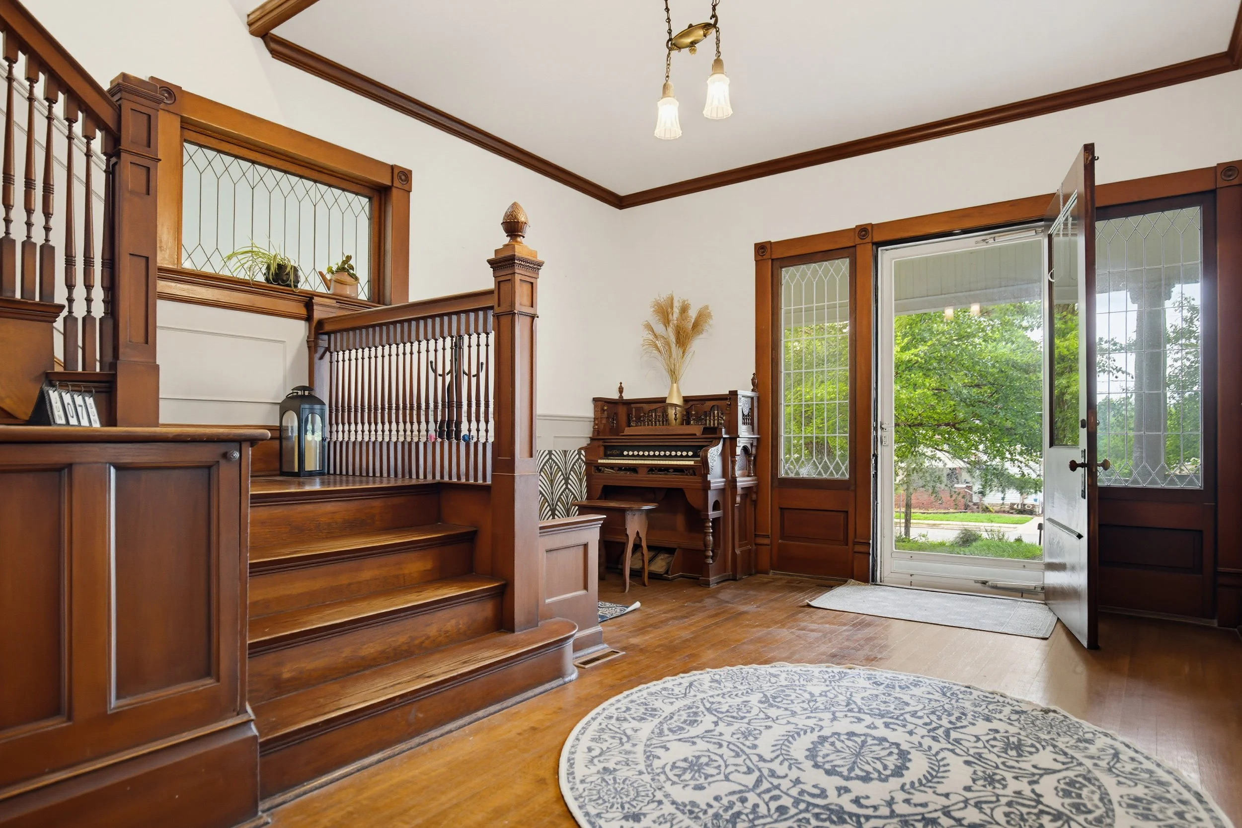 Living room with wooden staircase, open door with glass panels, and a view outside showing green trees.