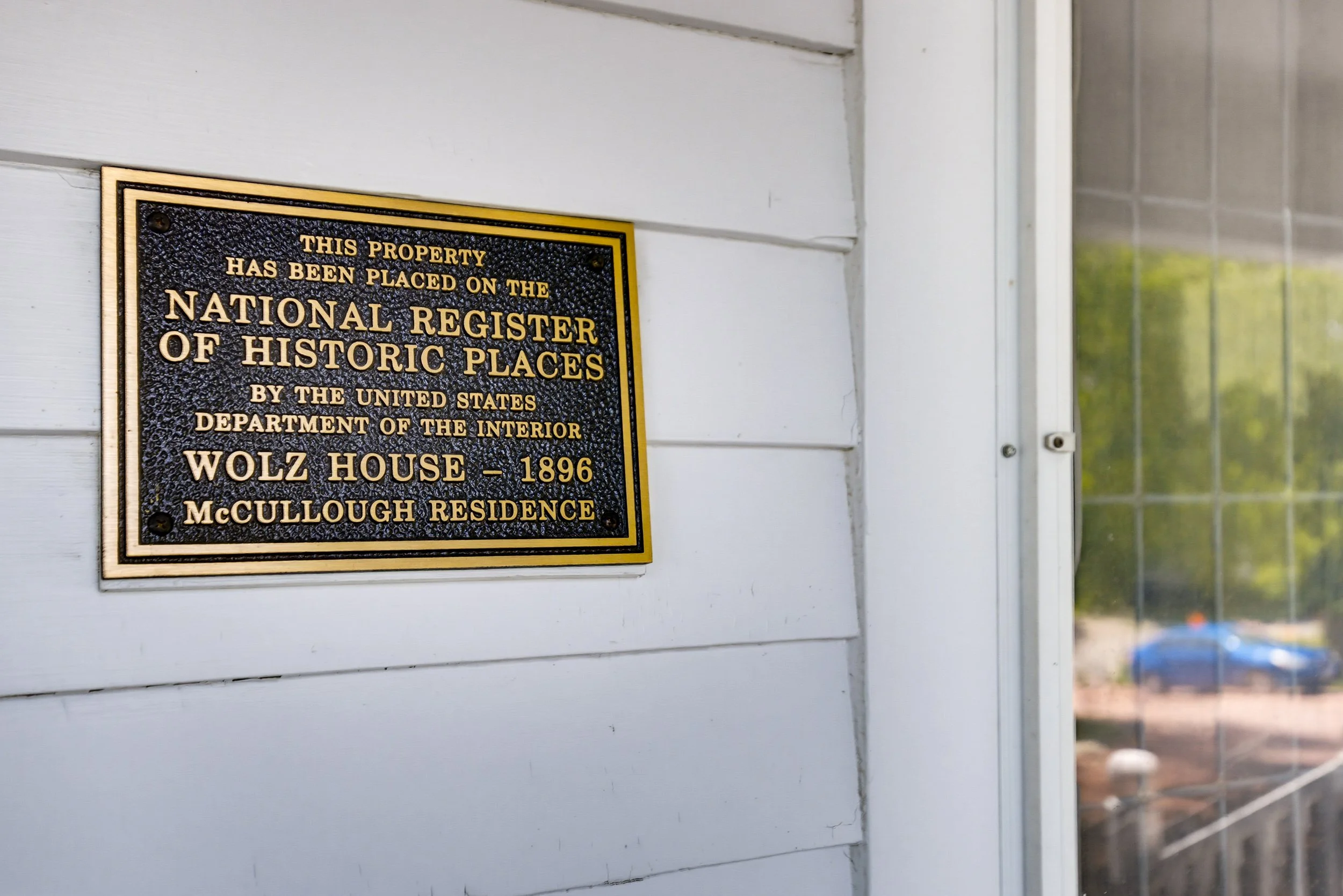 A bronze plaque mounted on white wooden siding on a house reads: