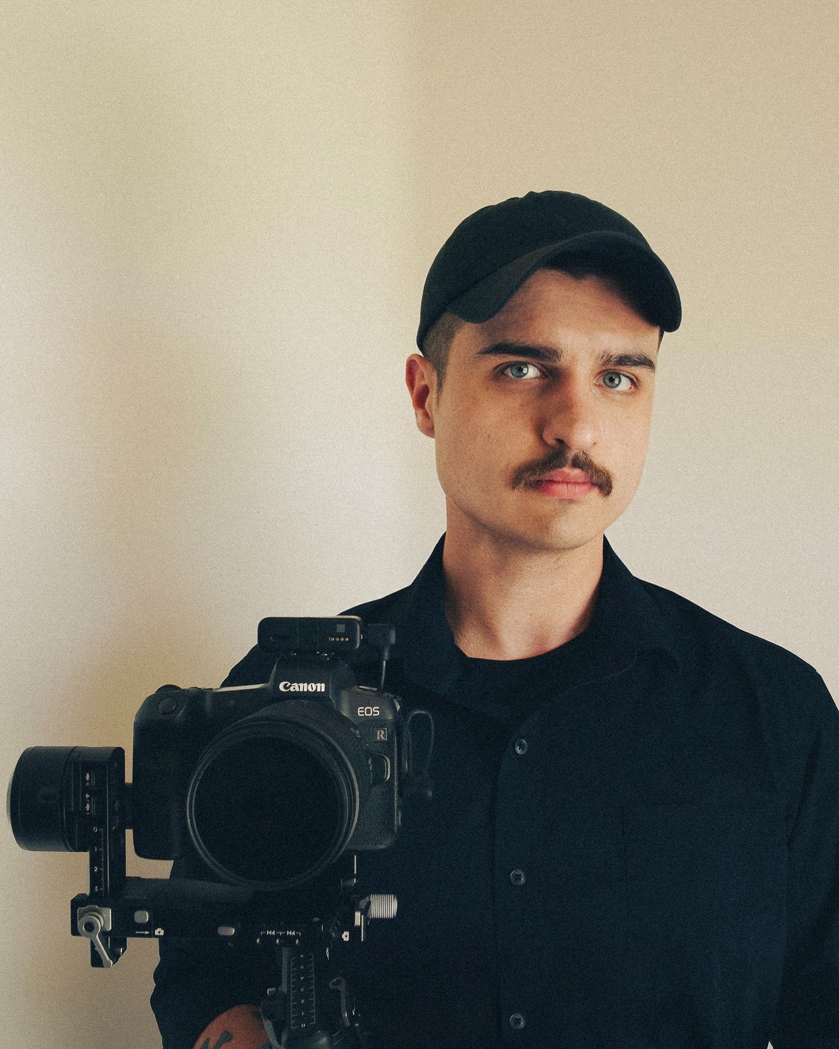 A young man with a mustache and blue eyes wearing a black cap and black shirt, holding a professional camera with a large lens.