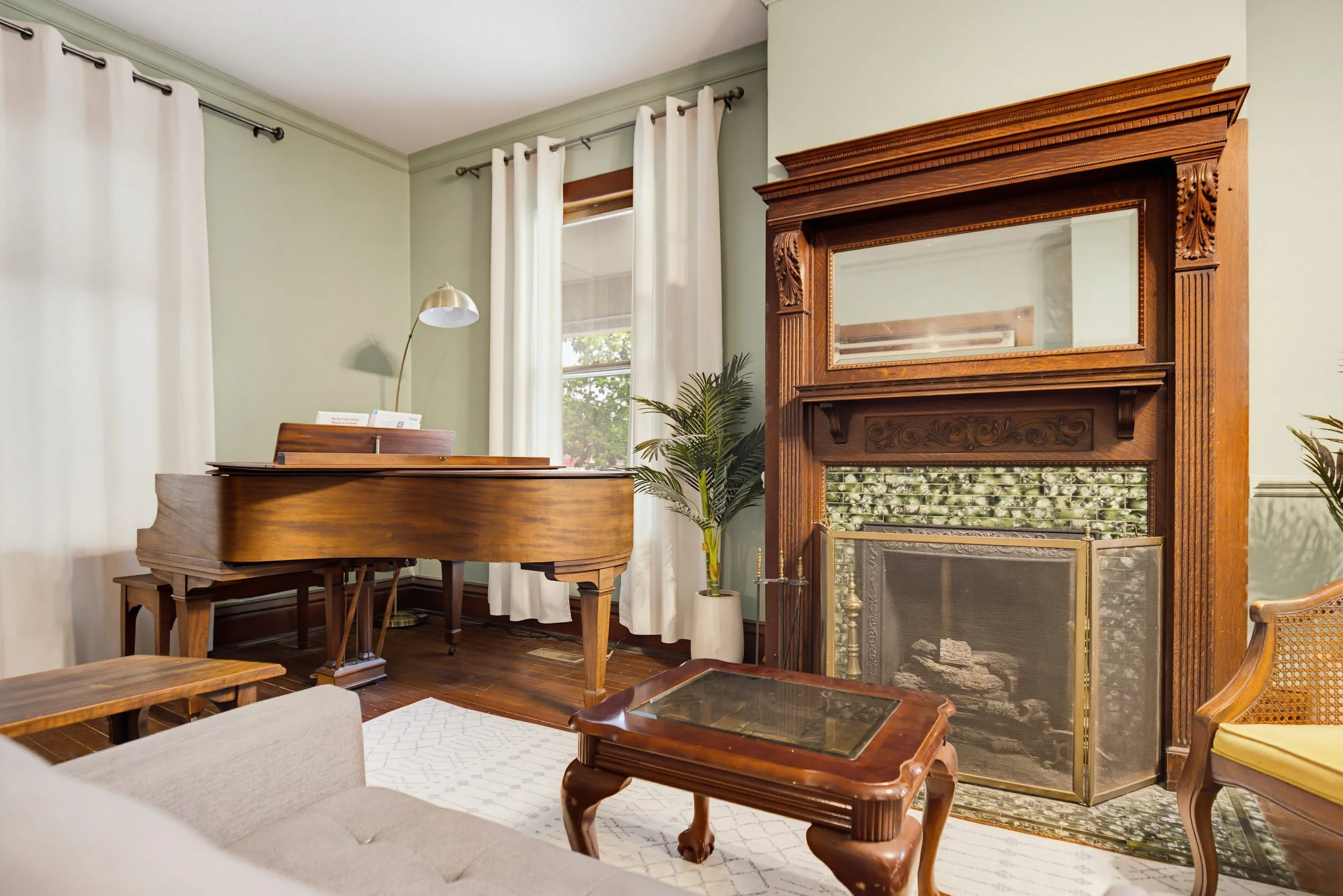 Living room with a wooden grand piano, window with white curtains, a fireplace with a large wooden mantel, a plant in a white pot, and seating furniture including a beige armchair and a wooden chair with a cane back.